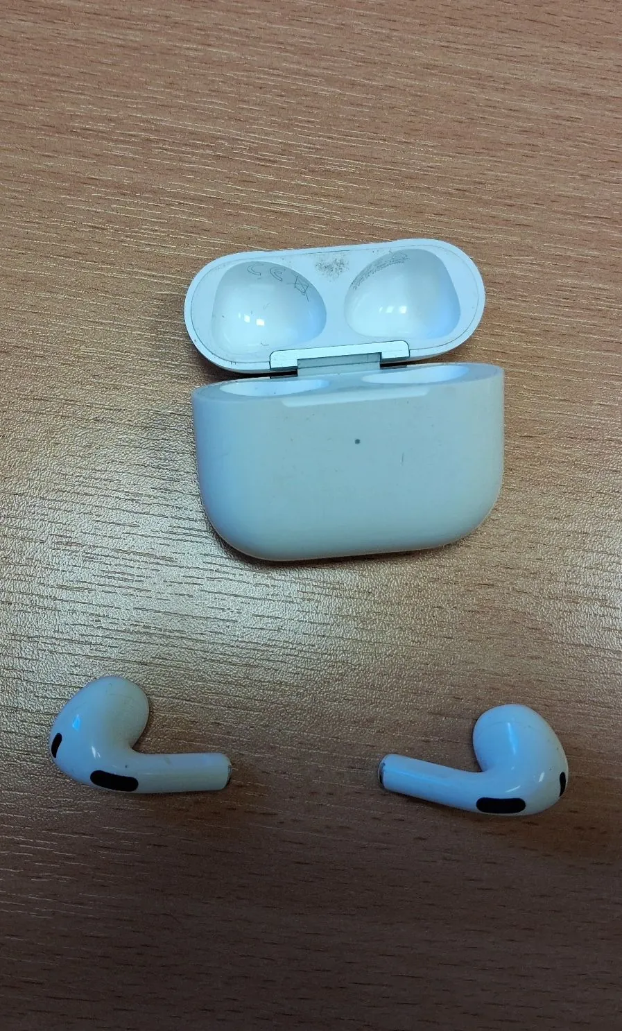 Białe słuchawki bezprzewodowe AirPods wraz z otwartym etui ładującym, leżące na drewnianej powierzchni o jasnobrązowym kolorze. Słuchawki znajdują się poniżej etui, oddzielone od siebie.