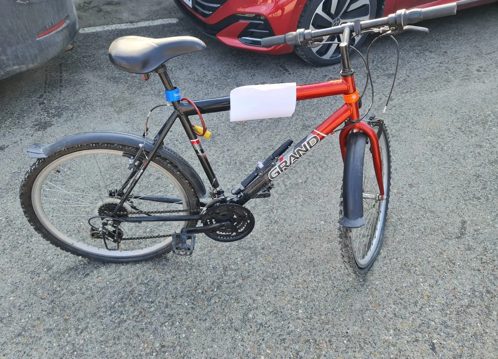 Czerwono-czarny rower marki GRAND z białym kartonem przytwierdzonym do ramy, stojący na asfaltowej powierzchni. W tle widoczne są częściowo dwa samochody: czerwony i szary. Rower posiada czarne koła, siedzisko oraz kierownicę, a na ramie widnieje napis GRAND. Na tle asfaltu z widocznymi liniami parkingu.