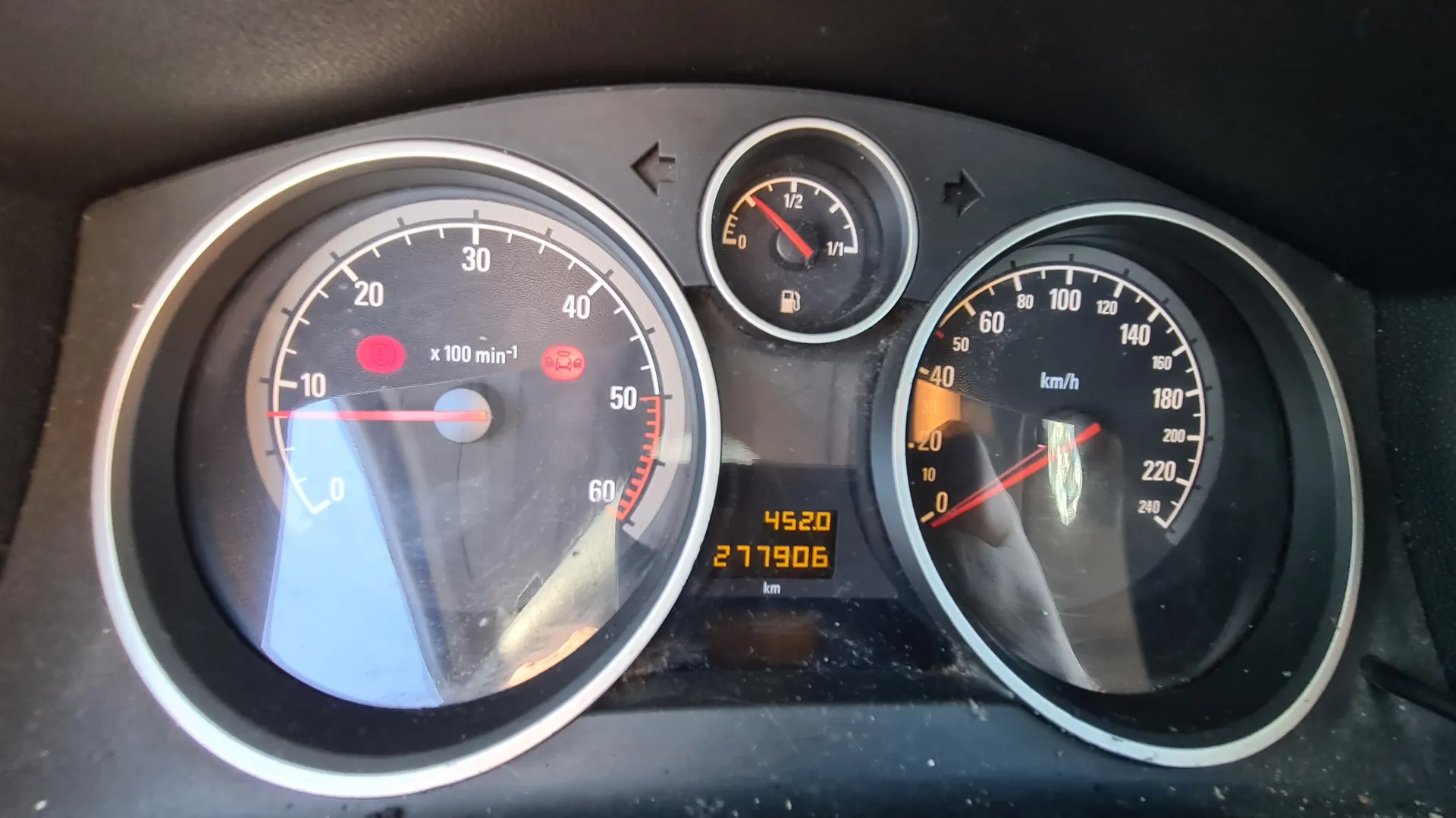 Tablica przyrządów samochodowych z trzema głównymi wskaźnikami. Po lewej stronie wskaźnik obrotomieru z zakresem od 0 do 60 (oznaczony jako x100 min⁻¹), z czerwonymi ikonami ostrzegawczymi. W środku mały wskaźnik paliwa z podziałką od 0 do 1/1 i ikoną zbiornika. Po prawej wskaźnik prędkości w km/h z zakresem do 240. Pomiędzy wskaźnikami wyświetlacz cyfrowy z licznikami: 4520 i 277906 km. Tło tablicy czarne, cyfry i znaczniki białe lub pomarańczowe, wskazówki czerwone. Widoczne odbicia na szybie przyrządów.