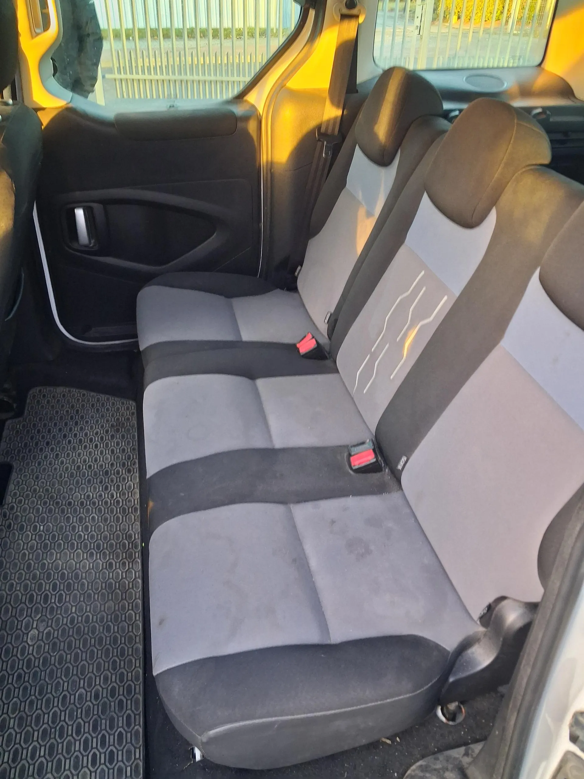 Wnętrze samochodu z widokiem na tylne siedzenia. Siedzenia są szaro-czarne z białymi liniami na oparciach. Widoczne są czerwone klamry pasów bezpieczeństwa. Drzwi samochodu są otwarte, a przez okno widać metalowe ogrodzenie i zieloną roślinność. Na podłodze czarny dywanik z sześciokątnym wzorem. Oświetlenie sugeruje poranek lub wieczór.