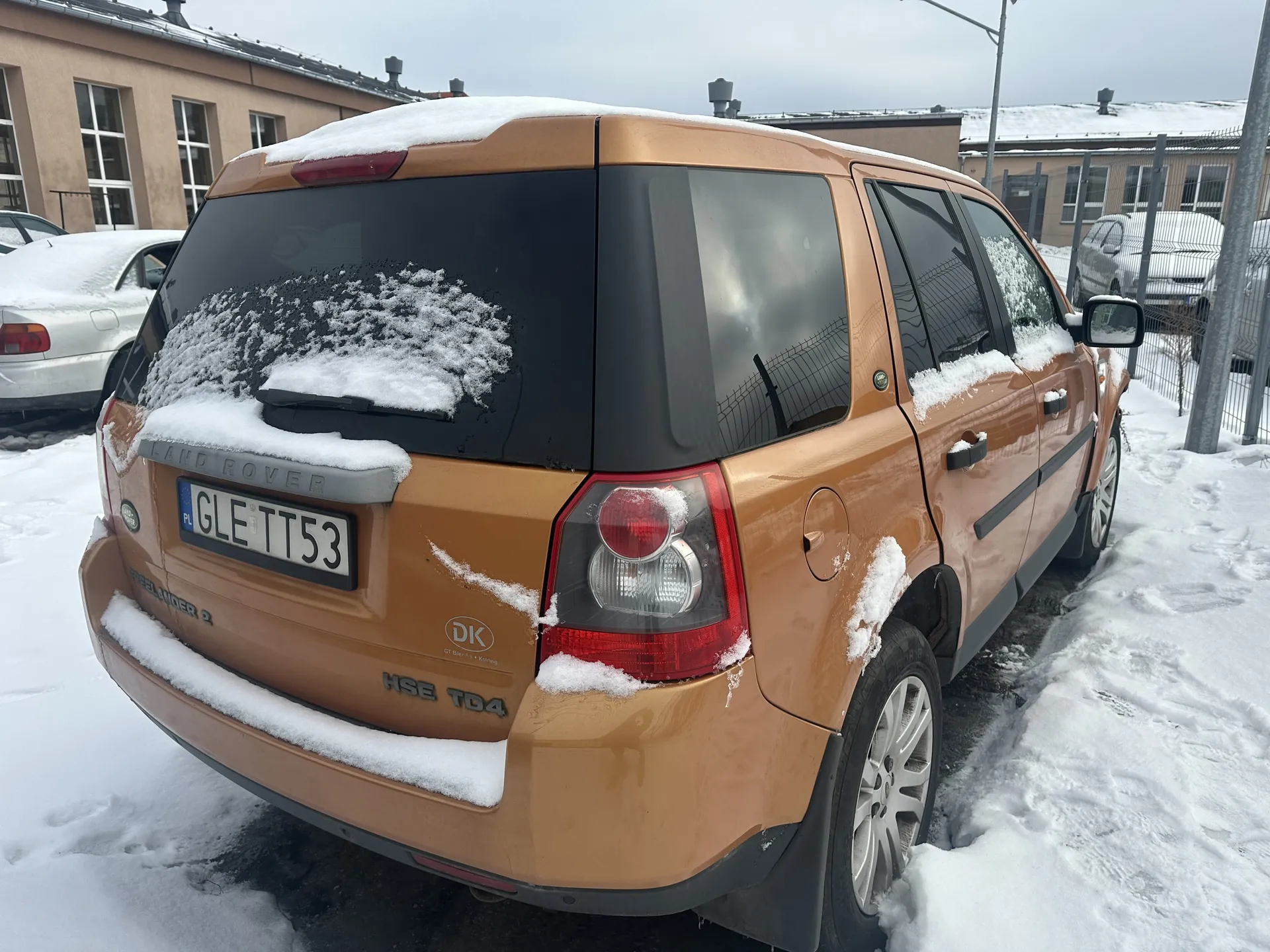 Zdjęcie przedstawia tył brązowego samochodu Land Rover Freelander 2 z oznaczeniem HSE TD4, zaparkowanego na śnieżnej powierzchni. Na dachu, szybie tylnej i części nadwozia widoczny jest śnieg. Tablica rejestracyjna PL GLE TT53 umieszczona jest pośrodku tyłu pojazdu. W tle znajdują się inne zaparkowane samochody, budynki o beżowych elewacjach oraz ogrodzenie. Niebo jest zachmurzone, a cała scena pokryta jest warstwą śniegu.