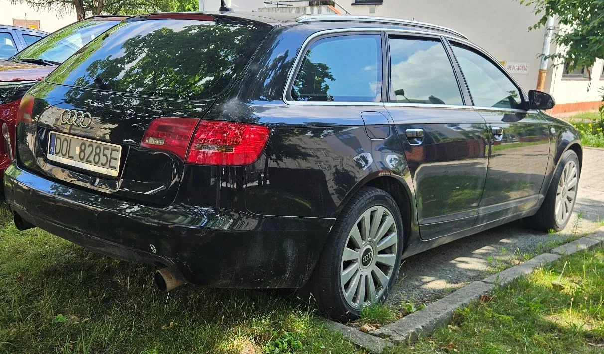 Czarny samochód marki Audi w nadwoziu kombi, zaparkowany na trawie obok krawężnika. Widoczny jest tył i prawy bok pojazdu. Samochód posiada polską tablicę rejestracyjną o numerze DOL 8285E.
