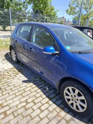 Niebieski samochód osobowy marki Volkswagen Golf zaparkowany na kostce brukowej. Widok z boku, z przodu i z góry. W tle znajduje się metalowy płot, zielone drzewa oraz fragment budynku i znak drogowy z literą P.