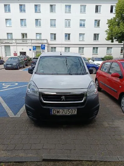 Srebrny samochód marki Peugeot zaparkowany przodem do obiektywu na brukowanym parkingu. Samochód ma tablicę rejestracyjną o numerze DW 7U507. W tle znajduje się biały, wielopiętrowy budynek z licznymi oknami oraz czerwony samochód zaparkowany po prawej stronie. Po lewej stronie widoczne jest niebieskie oznakowanie miejsca parkingowego dla osób niepełnosprawnych.