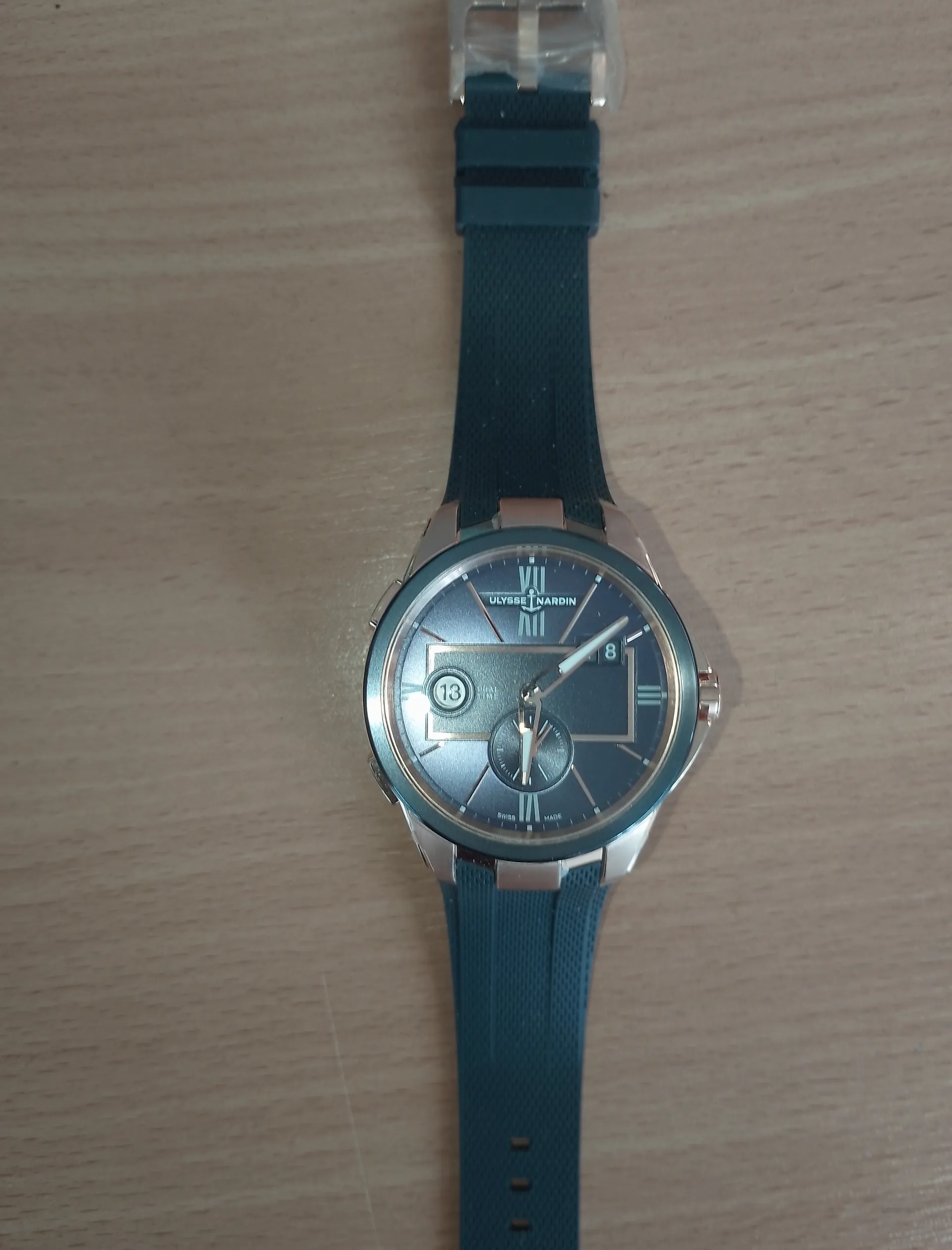 Zegarek Ulysse Nardin z ciemnoniebieską tarczą, rzymskimi cyframi i metalowym obwolutem. Tarcza zawiera okienka datowe z cyframi 13 i 8 oraz napisy 'ULYSSE NARDIN' i 'SWISS MADE'. Pasek zegarka jest ciemnoniebieski, wykonany z materiału przypominającego gumę. Zegarek leży na jasnobrązowej powierzchni drewnianej.