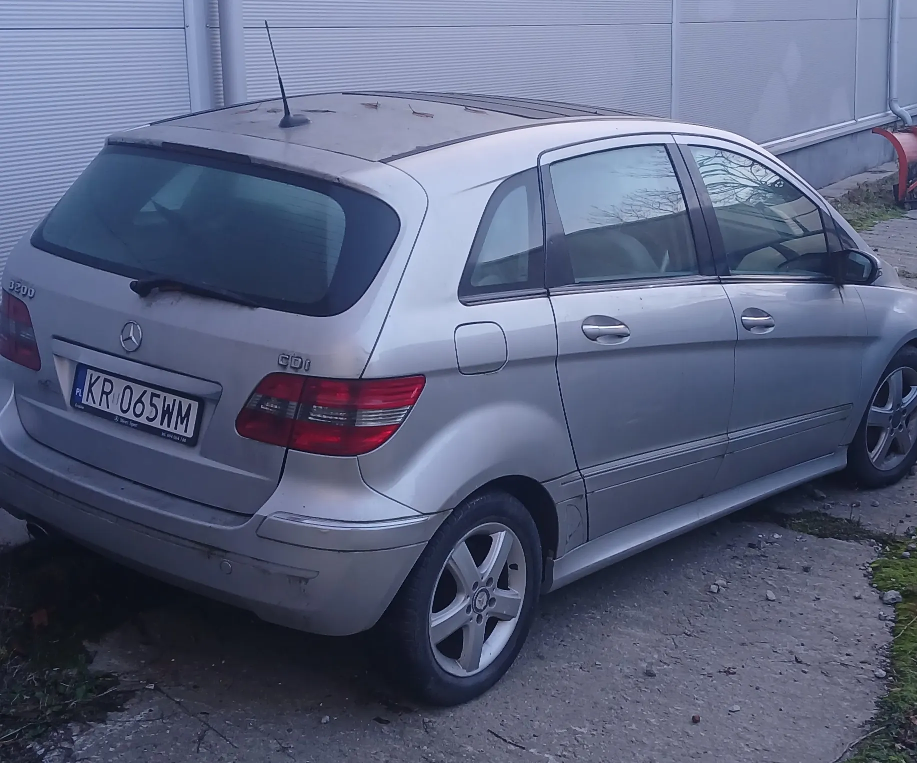 Srebrny samochód Mercedes-Benz klasy B, widok z tyłu i boku. Na tylnym zderzaku widoczny emblemat marki, napisy '3200' i 'CDI'. Tablica rejestracyjna z polskim kodem 'PL' i numerem 'KR 065WM'. Samochód zaparkowany na nierównym, żwirowym podłożu z fragmentami trawy. W tle szara, metalowa ściana budynku przemysłowego. Widoczne są czarne felgi, czerwone światła tylnego oraz antena na dachu.