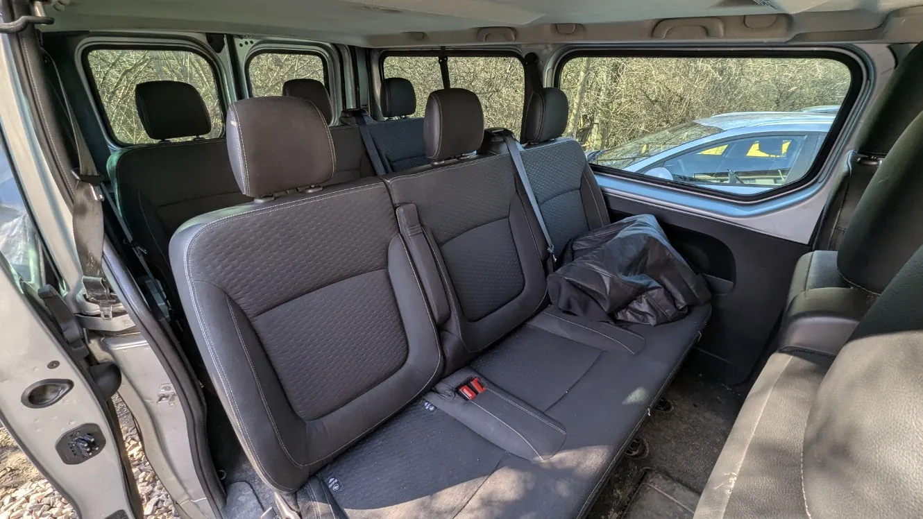Wnętrze furgonetki z czarnymi siedzeniami. Widoczne są trzy rzędy siedzeń, otwarte drzwi po lewej stronie, pasy bezpieczeństwa oraz czarna torba na jednym z siedzeń. Przez okna widać drzewa i częściowo inny samochód na zewnątrz. Wnętrze jest czyste, a siedzenia mają strukturalny wzór.