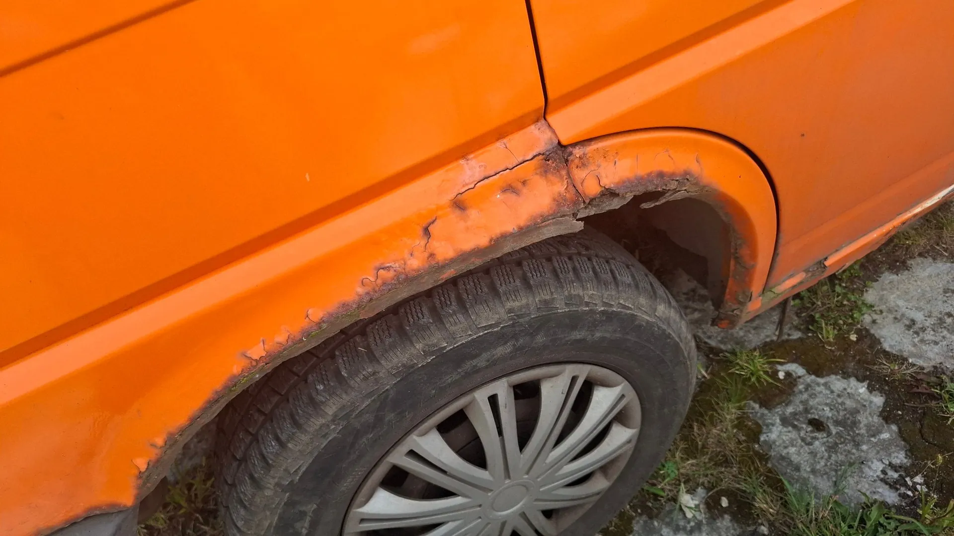 Zbliżenie na pomarańczowy element nadwozia samochodu nad przednim kołem. Metal wzdłuż nadkola jest silnie skorodowany, z widocznymi ubytkami w karoserii i łuszczącą się farbą. Widoczna jest część opony oraz srebrna felga z kołpakiem.