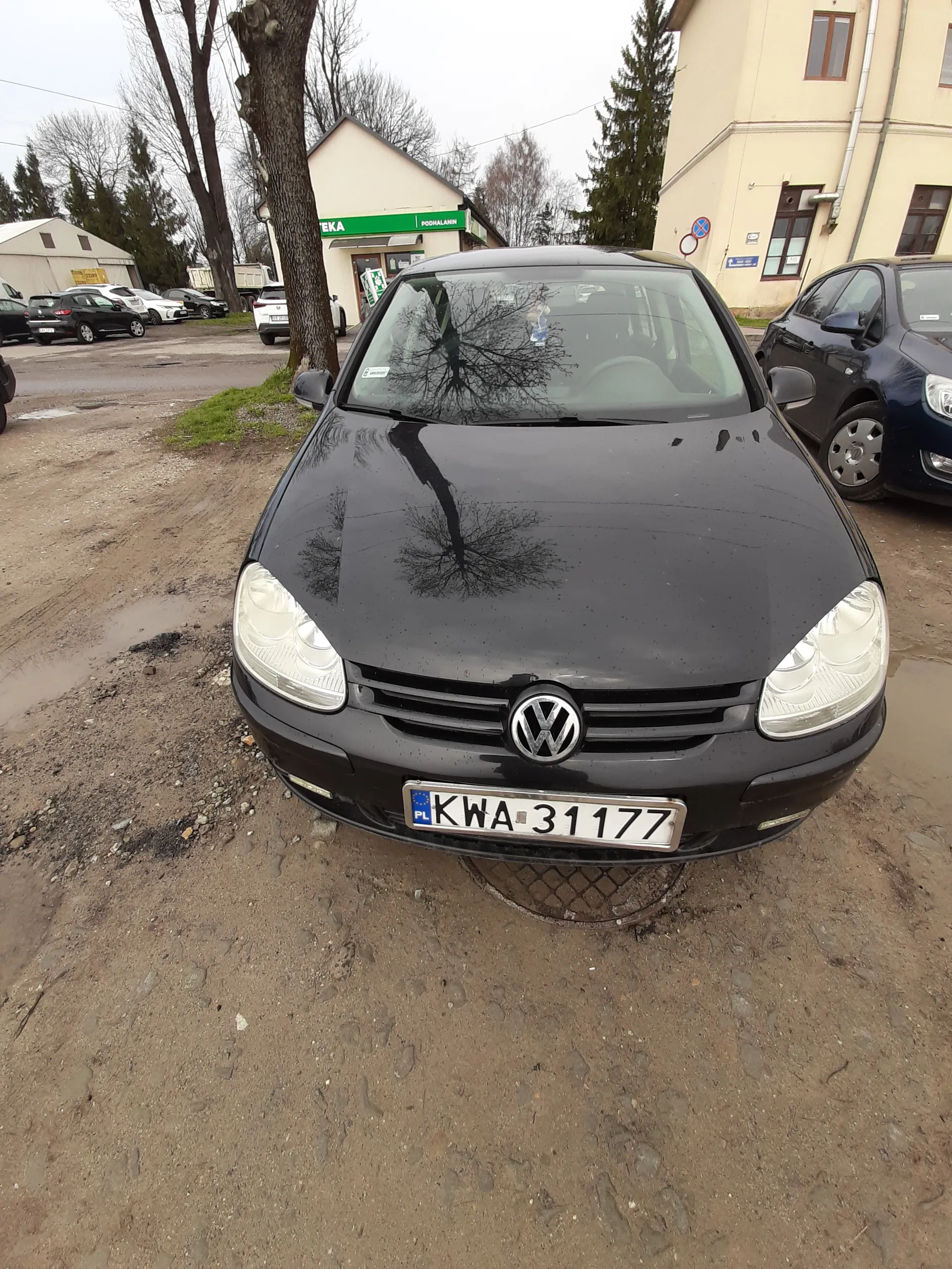 Czarny samochód marki Volkswagen z tablicą rejestracyjną KWA 31177 zaparkowany na brudnej, ziemistej powierzchni. W tle widoczne są inne pojazdy, budynki mieszkalne, drzewa bez liści oraz sklep z zielonym szyldem z napisem EKA. Na asfalcie znajdują się kałuże, a na tablicy rejestracyjnej widoczny jest znak PL. Odbicie drzewa na błyszczącej karoserii samochodu.