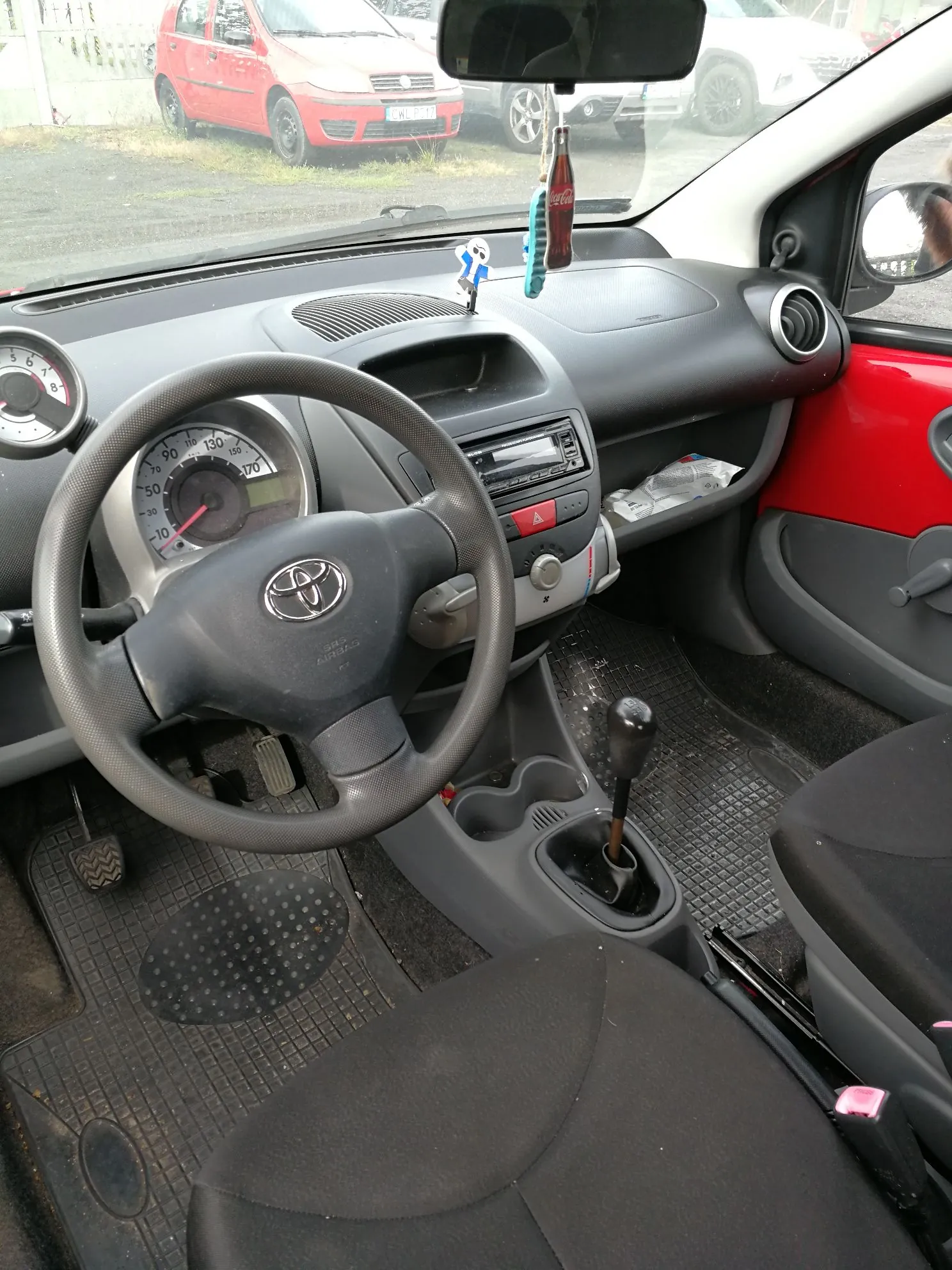 Wnętrze samochodu widziane z perspektywy kierowcy. Widoczna jest czarna kierownica z logo Toyoty, deska rozdzielcza z zegarami, radiem i nawiewami. Na lusterku wstecznym wisi butelka i niebieski brelok. W stopach znajdują się gumowe dywaniki. Przez przednią szybę widoczny jest czerwony samochód zaparkowany na zewnątrz.