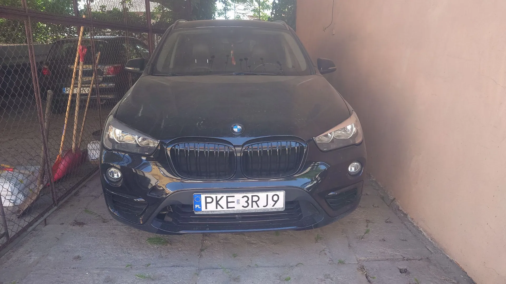 Czarny samochód marki BMW zaparkowany w garażu obok różowej ściany. Widoczny jest przód pojazdu z polską tablicą rejestracyjną o numerze PKE 3RJ9. W tle, za metalową bramą, stoi inny ciemny samochód oraz oparty o ogrodzenie drewniany drąg i czerwone narzędzie ogrodowe.