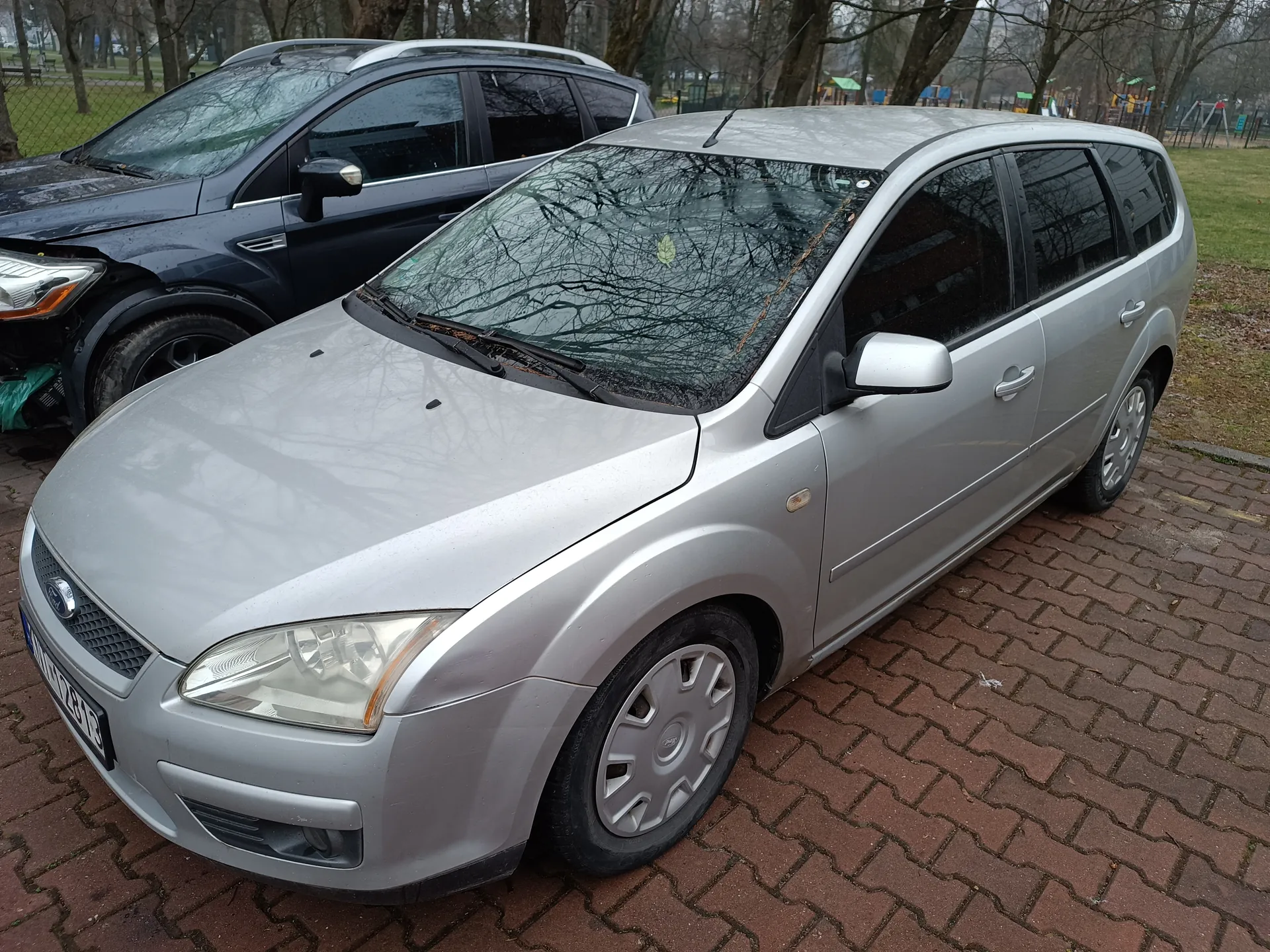 Srebrny samochód typu kombi marki Ford zaparkowany na ceglanym placu parkingowym. Widoczna tablica rejestracyjna WU 1234B. Za nim ciemnoszary SUV z uszkodzeniem przedniej lewej części. W tle drzewa bez liści, trawa i plac zabaw z kolorowymi elementami. Obraz jest prawidłowo zorientowany.