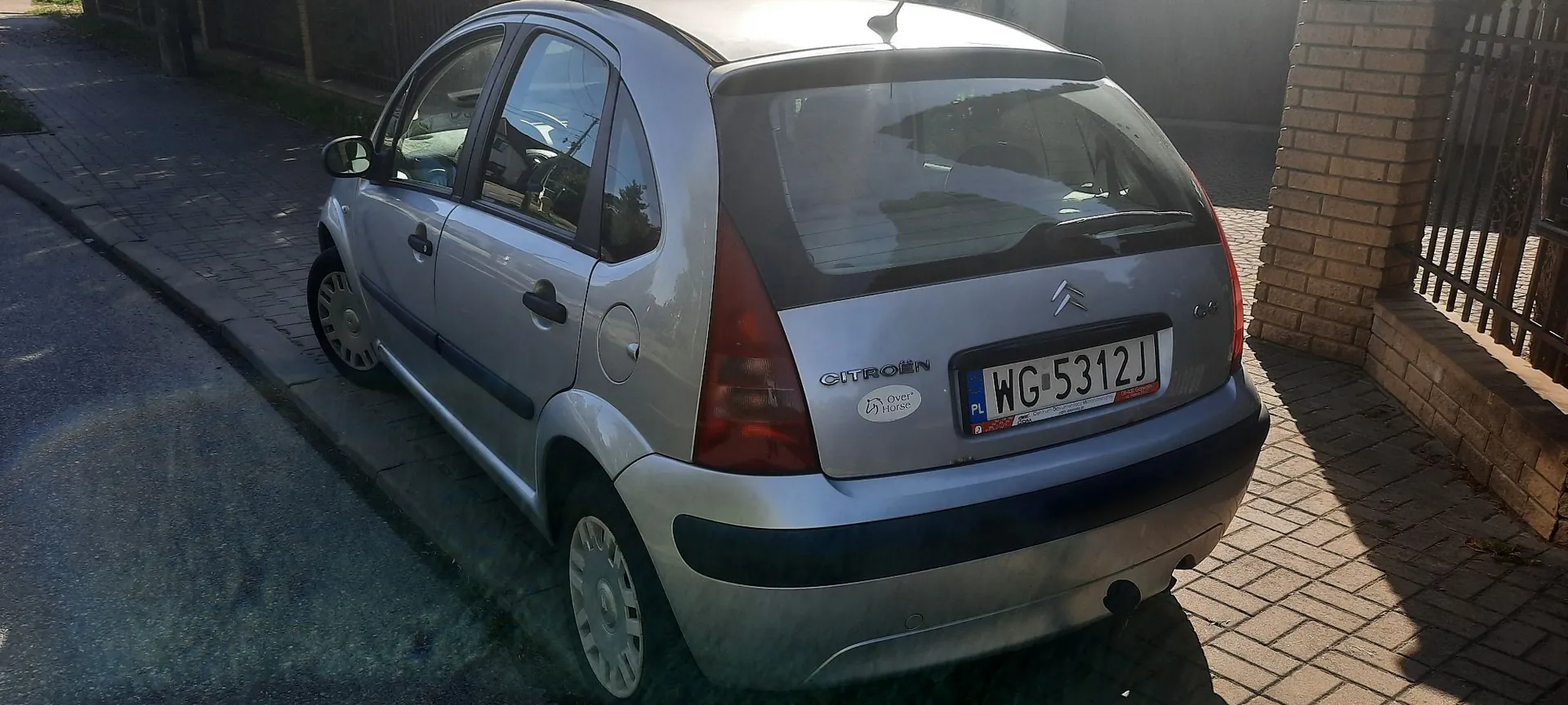 Srebrny samochód marki Citroën zaparkowany przy krawężniku na chodniku z kostki brukowej. Widoczny jest tył i lewy bok pojazdu. Na klapie bagażnika znajduje się logo Citroëna, napis CITROËN oraz polska tablica rejestracyjna o numerze WG 5312J.