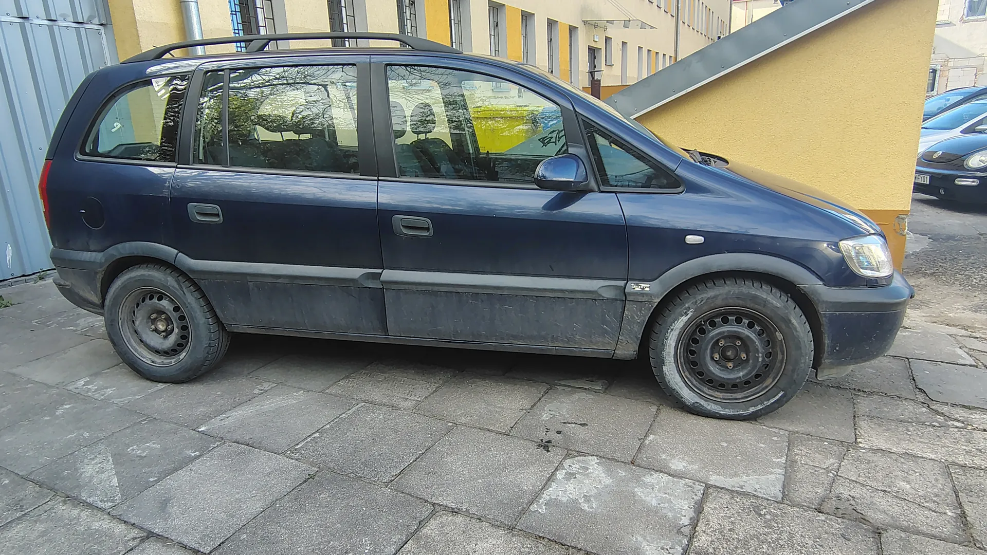 Ciemnoniebieski minivan zaparkowany na brukowanej powierzchni. Samochód ma zabrudzoną dolną część nadwozia, czarne felgi i dachowy bagażnik. W tle widoczny żółty budynek z oknami oraz fragment innych zaparkowanych pojazdów. Na przednim szybie minivana widoczne odbicia drzew i żółtego kontenera.