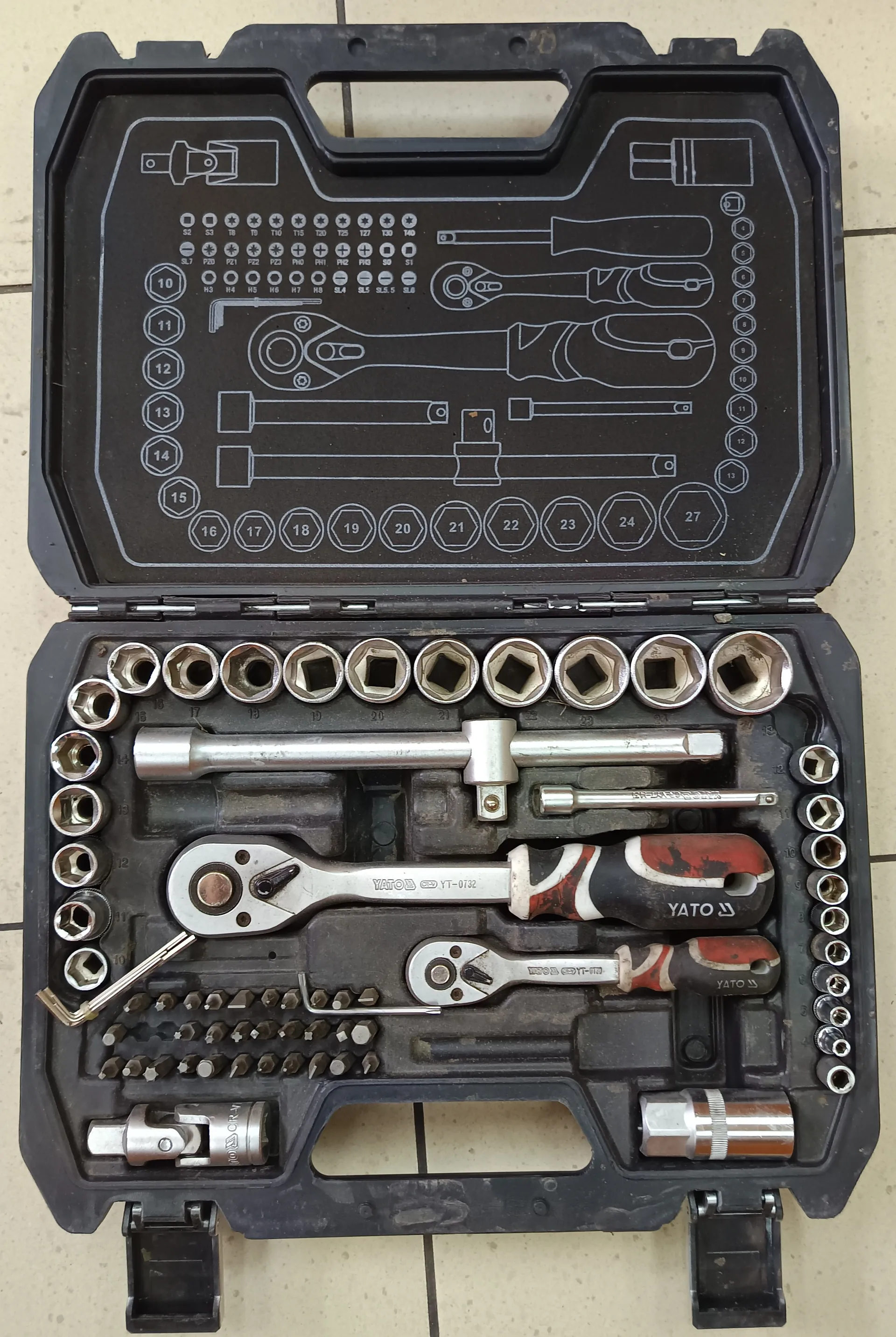 Otwarte czarne plastikowe etui narzędziowe z ułożonymi w nim narzędziami. W górnej części etui znajduje się schemat z oznaczeniami rozmiarów i miejscami na narzędzia, w tym cyfry od 10 do 27 oraz symbole gwintów. W dolnej części etui widoczne są metalowe nasadki, klucze nasadowe, dwa klucze z czerwono-czarnymi uchwytami z napisem YATO, długie metalowe rury oraz zestaw małych wkrętaków. Tło stanowi jasna płytkowa podłoga.