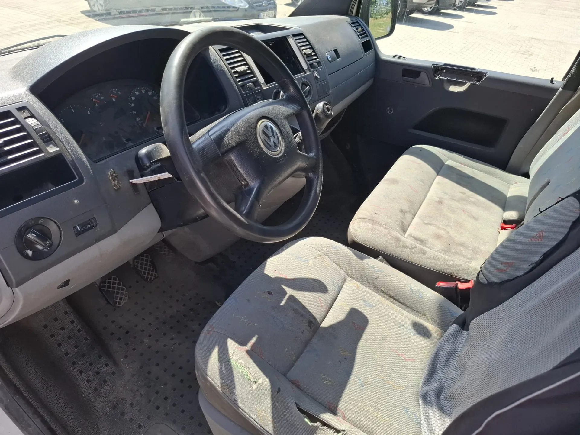 Wnętrze kabiny samochodu marki Volkswagen, widok z perspektywy pasażera. Widoczna jest kierownica z logo VW, deska rozdzielcza z zegarami, lewarek zmiany biegów oraz dwa szare, materiałowe fotele z widocznymi zabrudzeniami. Na podłodze znajdują się gumowe dywaniki i pedały.