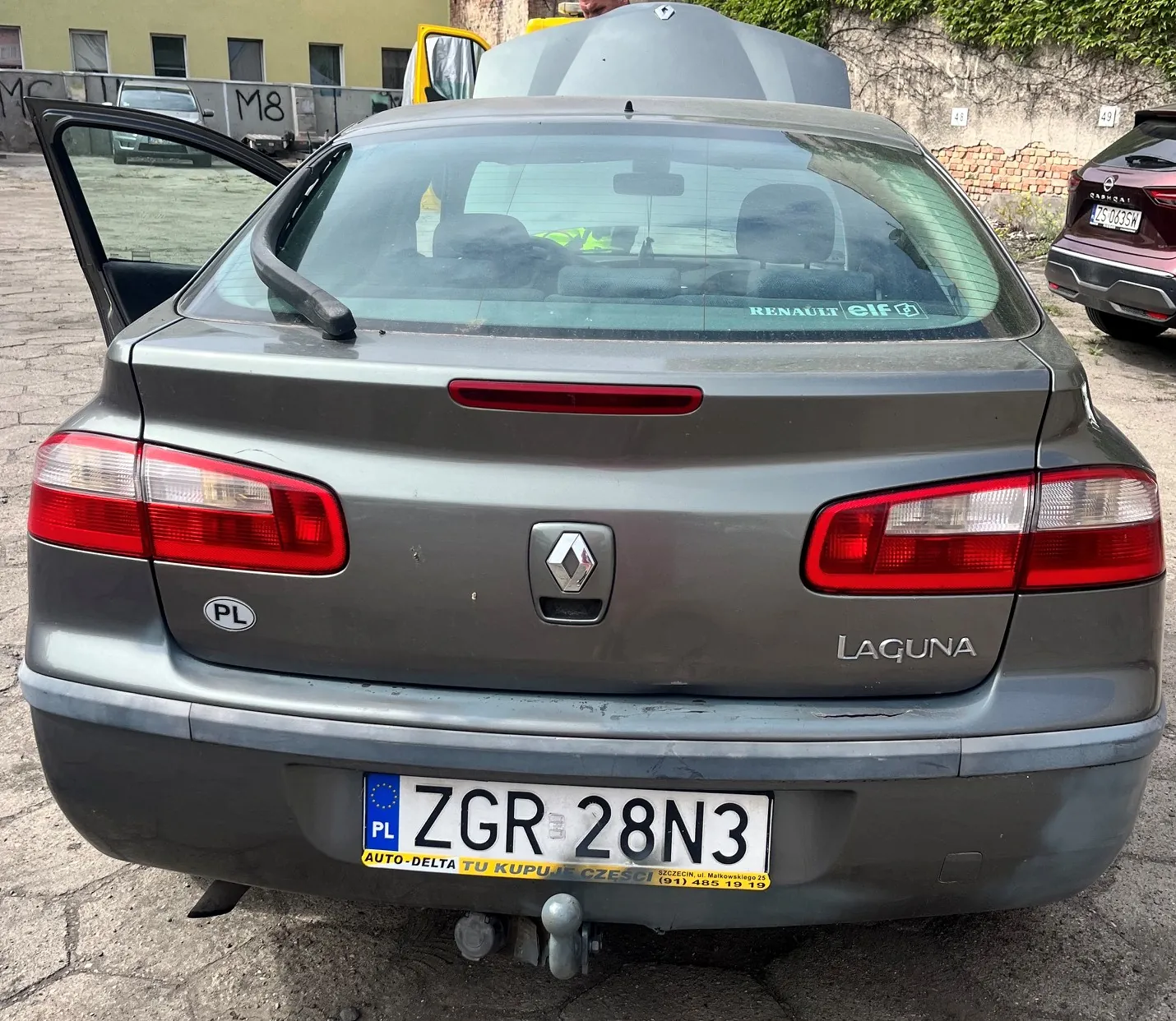 Tylny widok szarego samochodu marki Renault Laguna. Pojazd ma otwartą lewą tylną stronę oraz otwartą klapę bagażnika. Na klapie znajduje się logo Renault oraz napis 'LAGUNA'. Samochód posiada polską tablicę rejestracyjną o numerze ZGR 28N3 z żółtym paskiem reklamowym 'AUTO-DELTA TU KUPUJĘ CZĘŚCI' oraz numerem telefonu (91) 485 19 19. Po lewej stronie widoczna jest naklejka 'PL'.