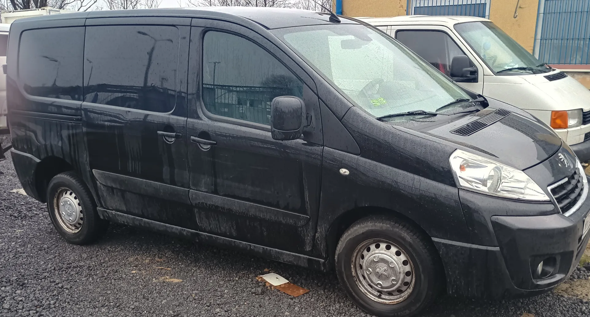 Zdjęcie przedstawia czarny furgon marki Peugeot zaparkowany na podwórku z żwiru. Furgon jest zabrudzony, z widocznymi plamami błota na bokach i przedniej części. W tle znajduje się biały furgon oraz budynek z niebieską bramą i bezlistnymi drzewami. Na przedniej szybie czarnego furgona widoczny jest żółty znaczek. Podwozie i koła furgona mają ślady zużycia. Na żwirze leży kawałek kartonu lub podobnego materiału.