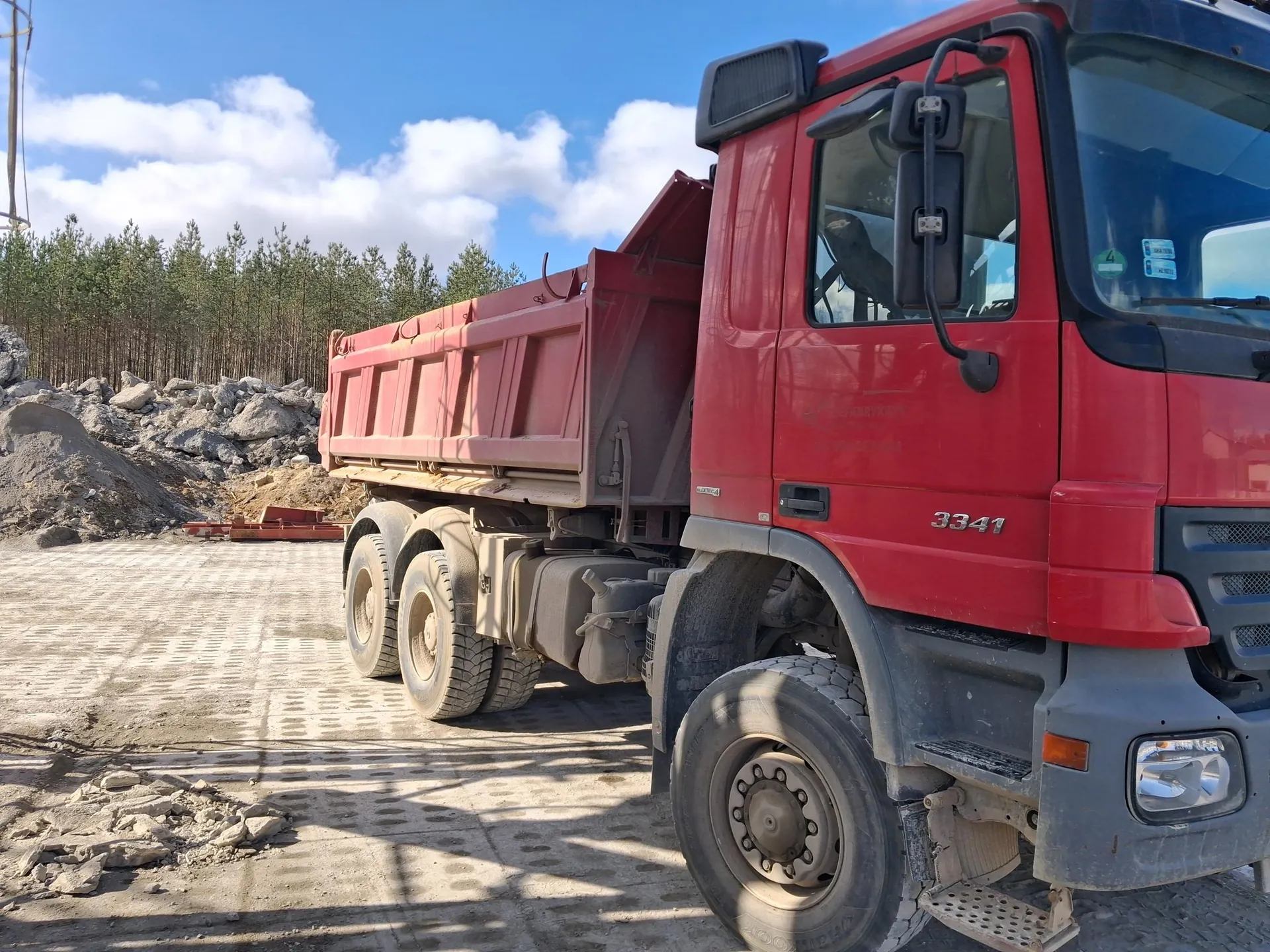 Czerwony samochód ciężarowy typu wywrotka z numerem 3341 na drzwiach kierowcy. Pojazd stoi na betonowej powierzchni z widocznymi śladami opon i rozsypanymi kamieniami. W tle stosy skał, gęsty las iglasty oraz niebo z białymi chmurami. Na przedniej szybie widoczne są naklejki, a nad drzwiami umieszczono czarny wentylator. Opony i elementy podwozia pokryte są kurzem.
