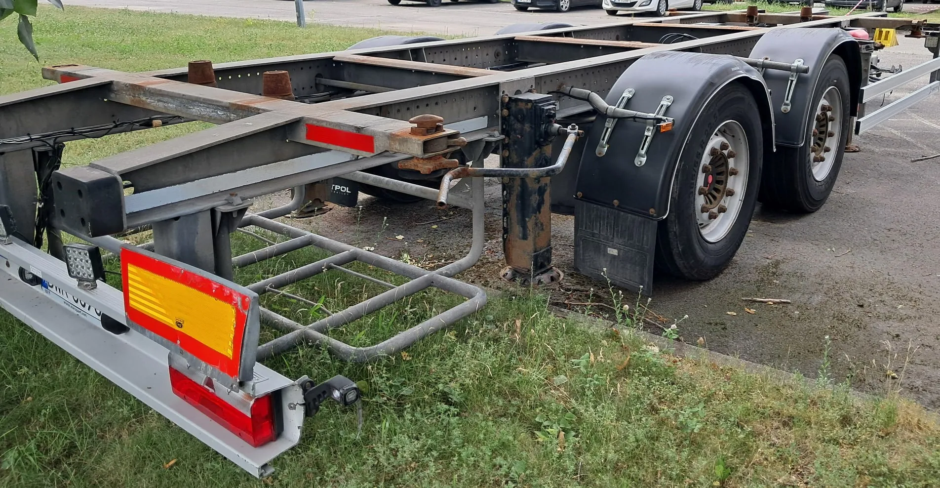 Zdjęcie przedstawiające metalową ramę przyczepy transportowej z dwiema osiami i kołami. Rama jest szara, z widocznymi elementami konstrukcyjnymi, wspornikami oraz czerwono-żółtymi odblaskami z tyłu. Koła są osłonięte czarnymi błotnikami. Przyczepa stoi na trawiastym poboczu obok asfaltowej drogi.