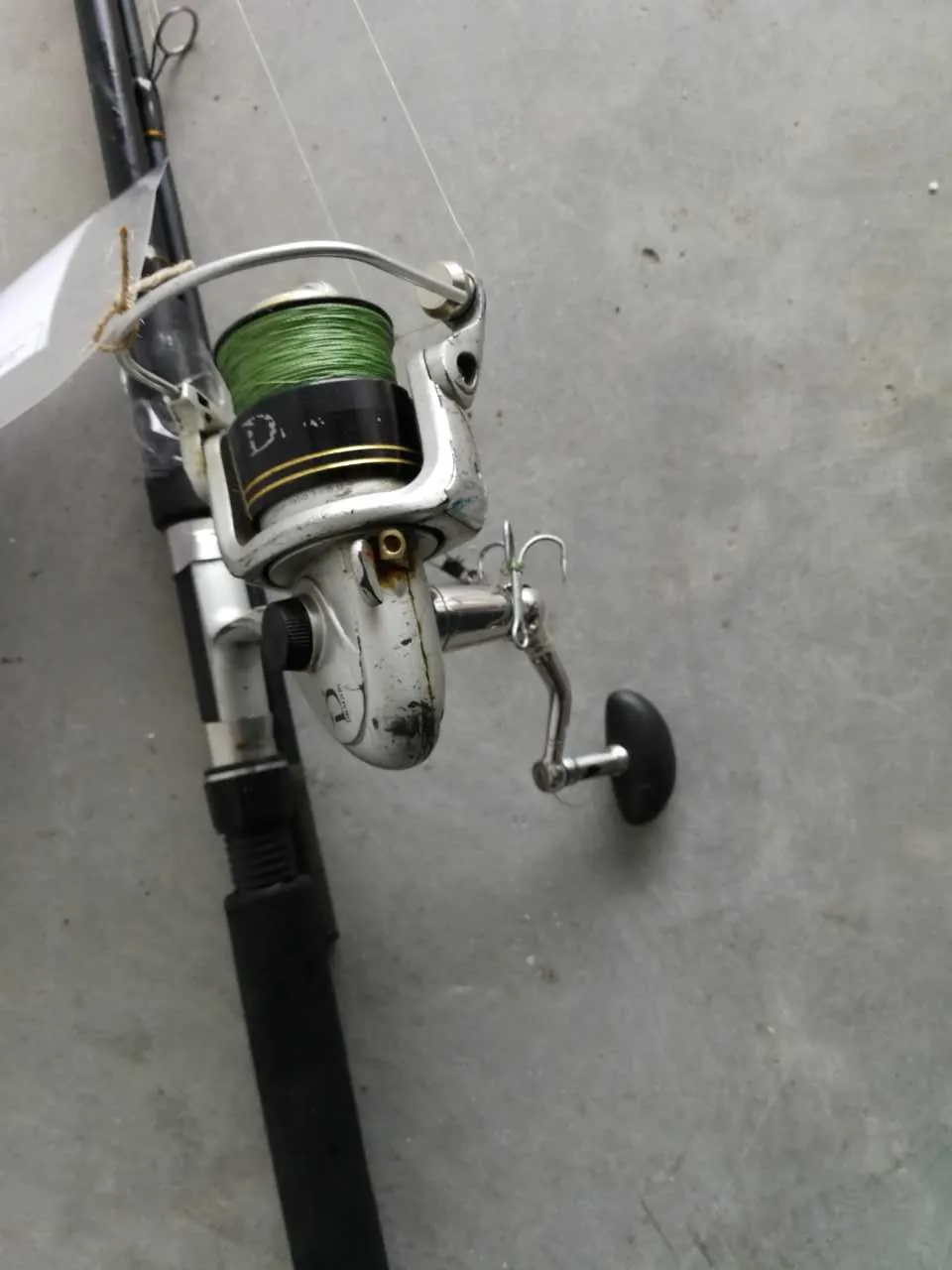 Wędkarska kołowrotka z zielonym sznurem zamocowana na czarnym wędce. Kołowrotka posiada metalowe elementy z widocznym napisem '10' oraz czarną rączkę. W tle szara powierzchnia betonowa. Do wędki przyczepiony jest biały etykietka z nieczytelnym tekstem.