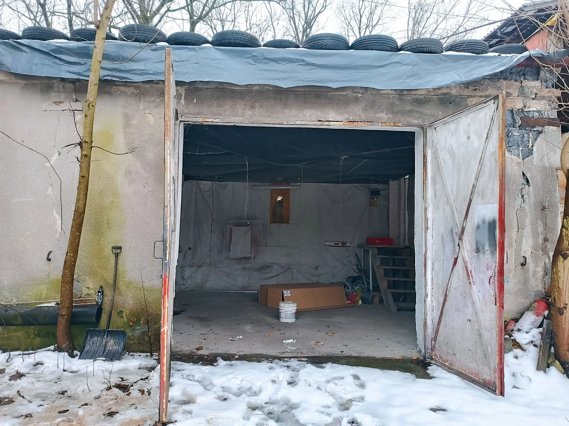 Zdjęcie starego, betonowego garażu z otwartymi, metalowymi drzwiami. Dach garażu jest przykryty szarą plandeką, na której ułożono w rzędzie czarne opony samochodowe. Wewnątrz garażu widać białą plandekę na ścianach, kartonowe pudełko na podłodze oraz drewnianą konstrukcję przypominającą schody lub regał. Przed garażem leży śnieg, a po lewej stronie stoi łopata oparta o ścianę obok cienkiego drzewa.