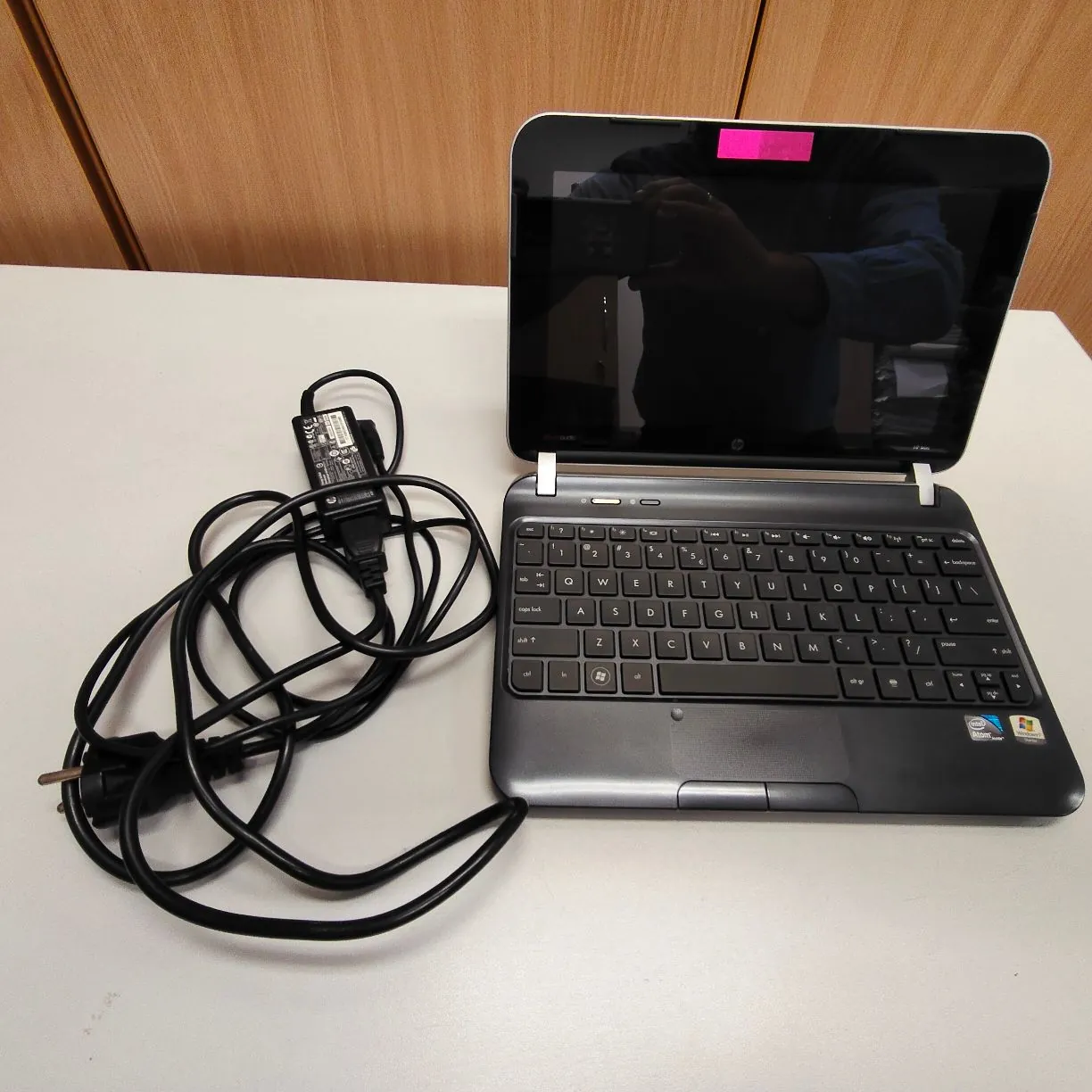 Srebrny laptop marki HP z otwartym ekranem, stojący na białym blacie. Obok laptopa znajduje się czarny zasilacz z długim kablem i wtyczką sieciową. Na ekranie laptopa widoczne jest odbicie osoby robiącej zdjęcie oraz mała, różowa naklejka w górnej części ramki.