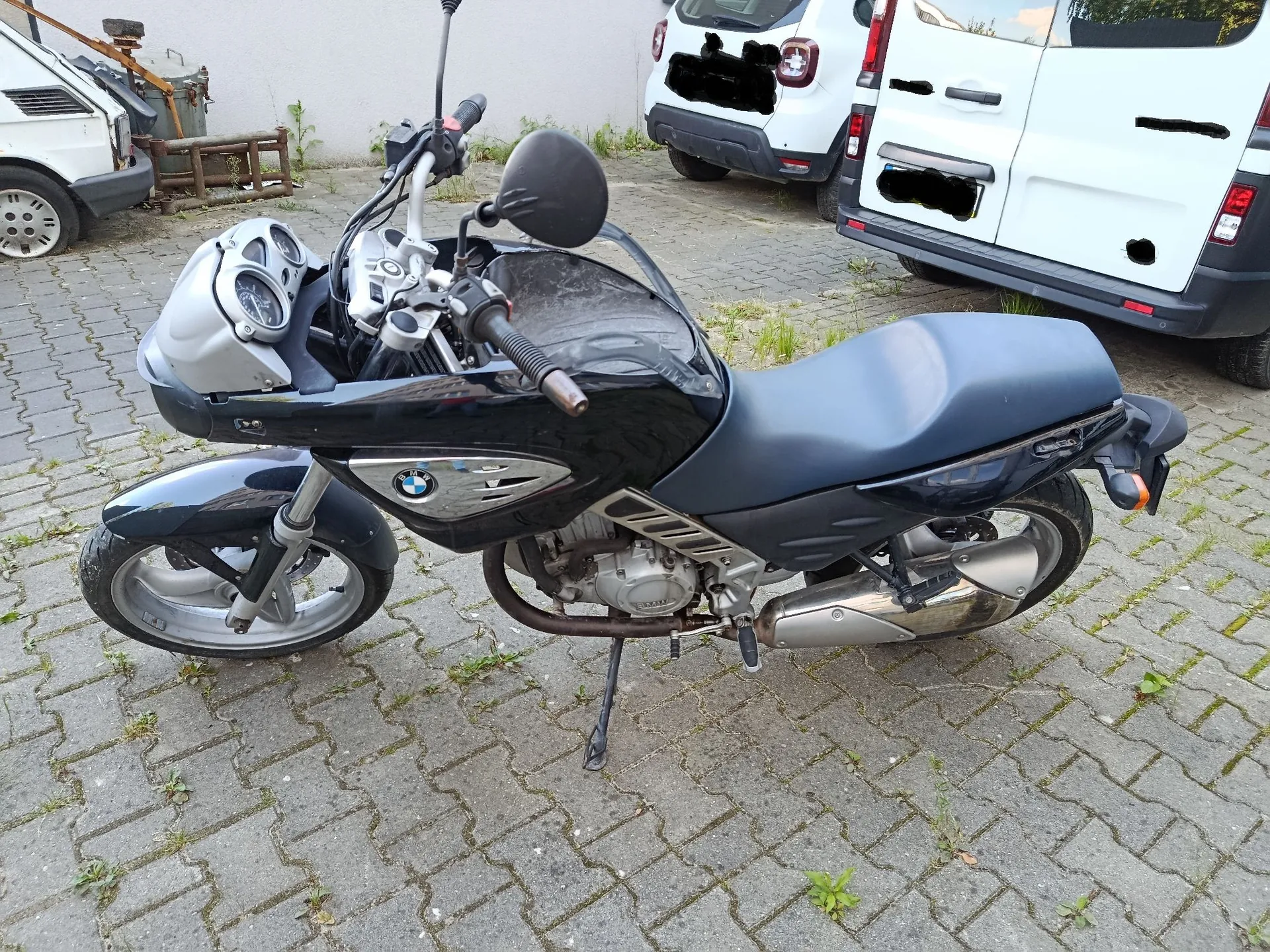 Czarny motocykl marki BMW zaparkowany na kostce brukowej. Motocykl stoi na bocznej stopce, widoczny jest z profilu. W tle znajdują się białe samochody oraz fragmenty infrastruktury miejskiej.