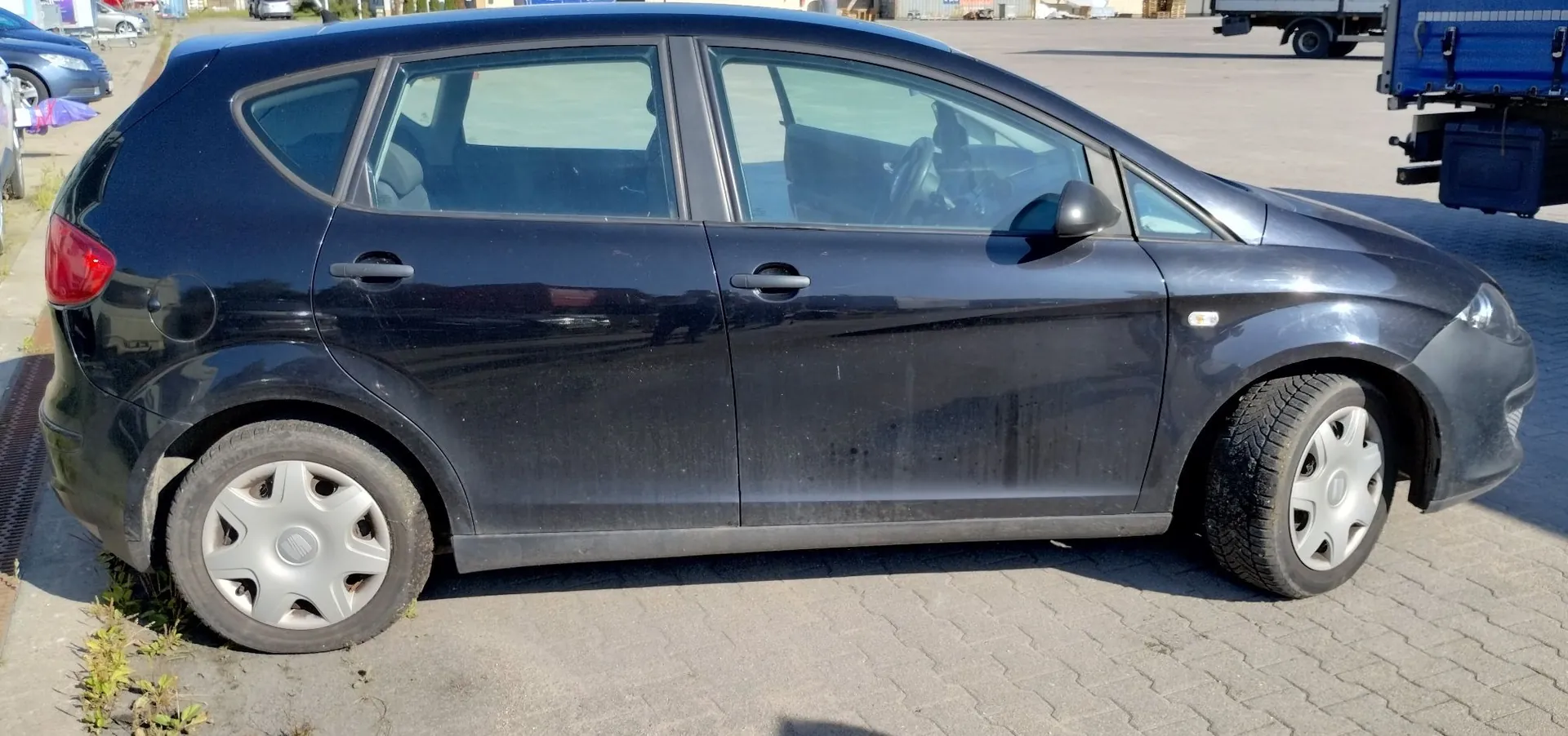 Czarny samochód osobowy typu hatchback zaparkowany bokiem na kostce brukowej. Pojazd jest zakurzony, posiada srebrne kołpaki na kołach. W tle widoczny fragment innego niebieskiego samochodu oraz ciężarówki.