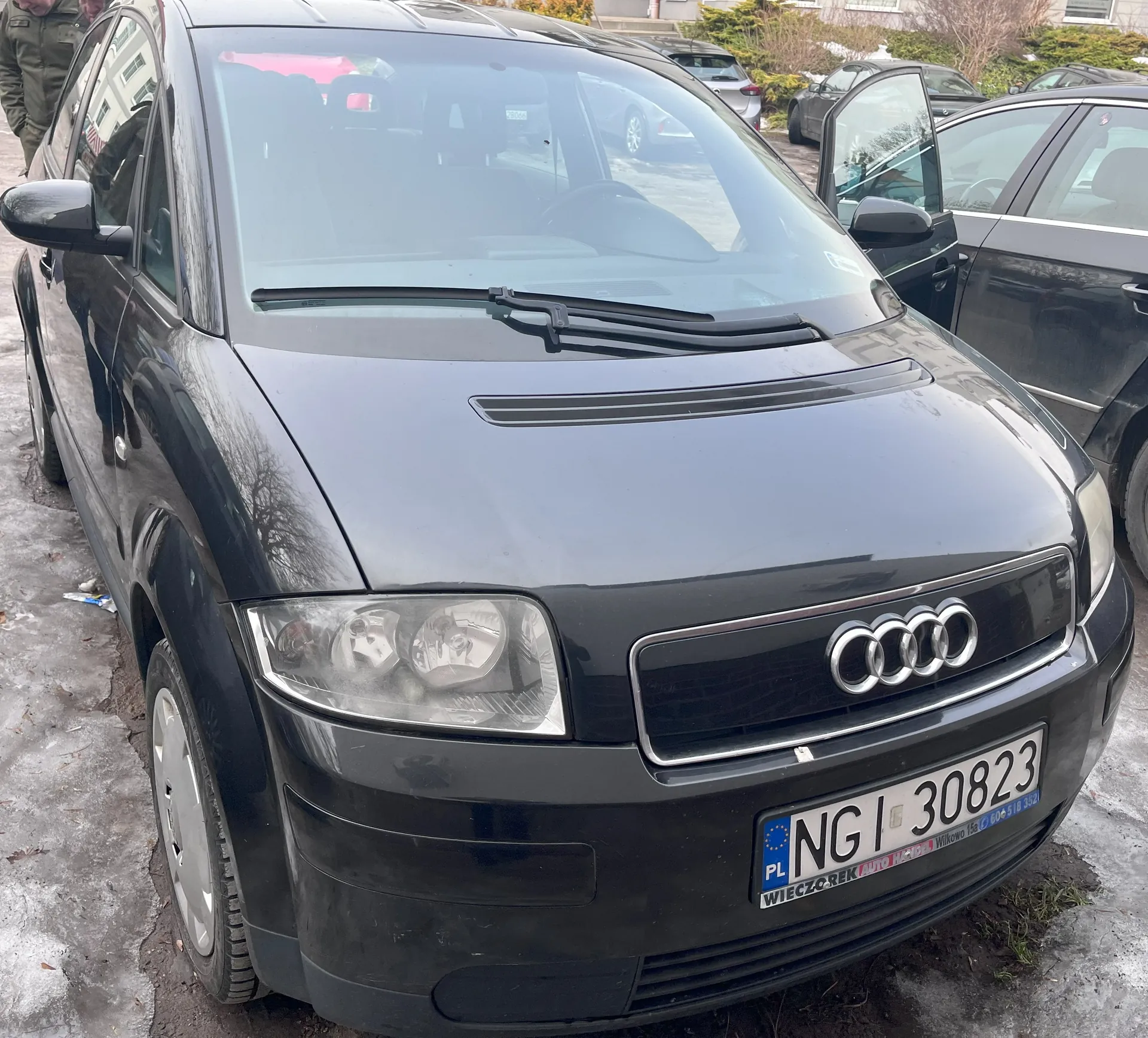 Czarny samochód marki Audi z przodu, z widocznym logo na atrapie chłodnicy. Tablica rejestracyjna z polskim oznaczeniem PL, numerem NGI 30823 oraz dodatkowym napisem WIECZÓR EK AUTO HACZEK i numerem telefonu 604 518 352. Samochód zaparkowany na zamarzniętej nawierzchni, w tle inne pojazdy, w tym jeden z otwartymi drzwiami. Po lewej stronie widoczna osoba w zielonej kurtce. W tle budynki i krzewy.
