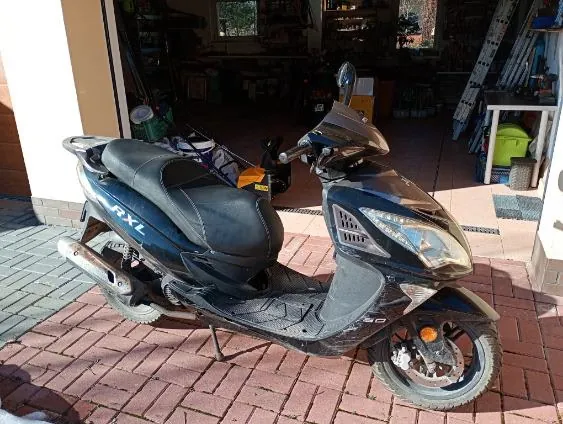 Czarny skuter marki Honda RFXL zaparkowany na ceglanym chodniku przed otwartym garażem. W tle widoczne narzędzia, pojemniki i sprzęt wewnątrz garażu. Skuter ma czarne siedzenie, błyszczące elementy i widoczny napis 'RFXL' na boku. Oświetlenie słoneczne rzuca cienie na powierzchnię.