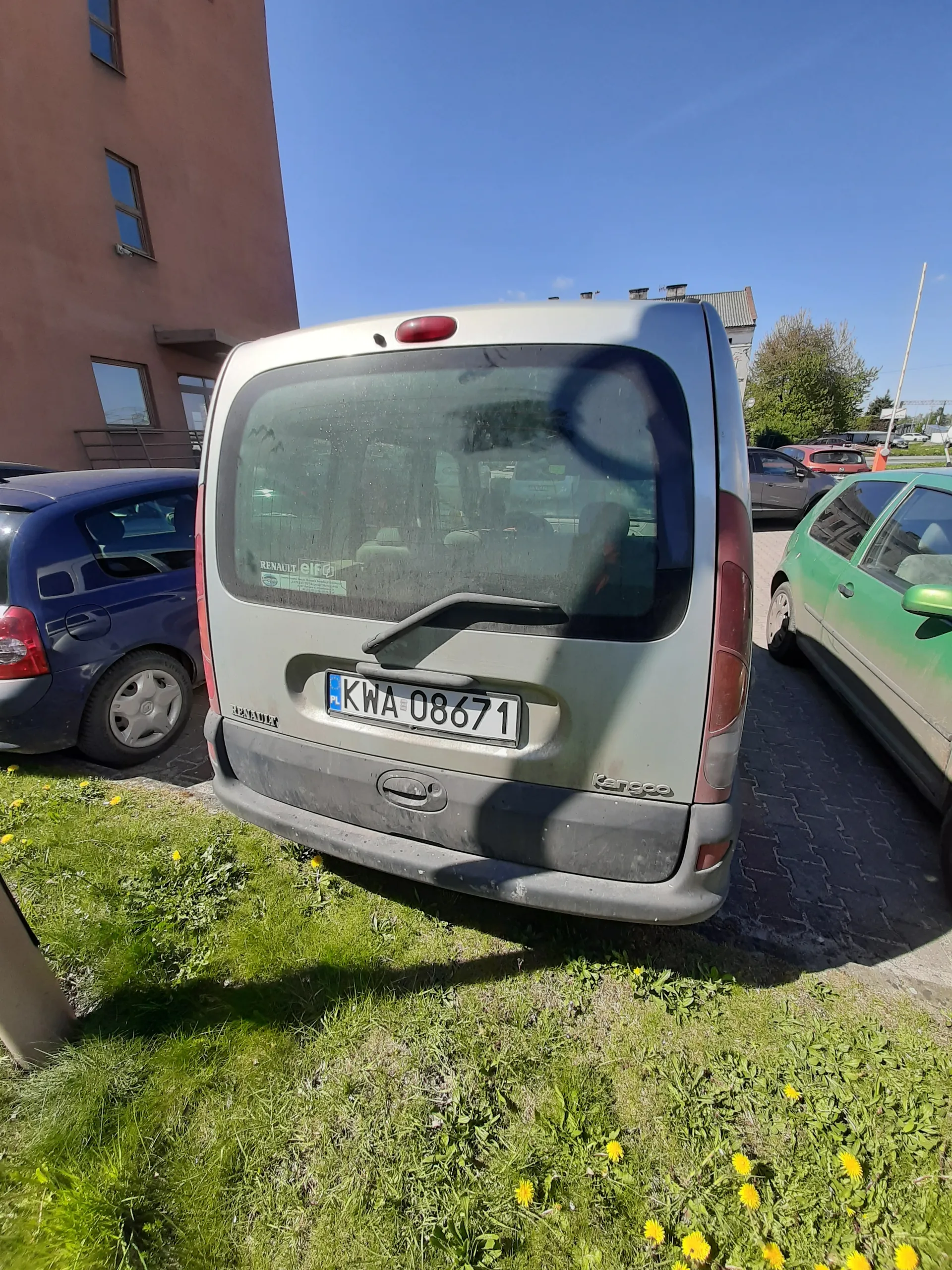 Zdjęcie przedstawia srebrny samochód Renault Kangoo z tablicą rejestracyjną KWA 08671, zaparkowany na brukowanej powierzchni. W tle widoczne są inne pojazdy: zielony samochód i niebieski samochód. Po prawej stronie znajduje się trawnik z żółtymi kwiatami (mniszkiem lekarskim). W tle również brązowy budynek z oknami oraz jasne niebo. Tablica rejestracyjna jest pionowo ustawiona, co wskazuje na nieprawidłową orientację zdjęcia.