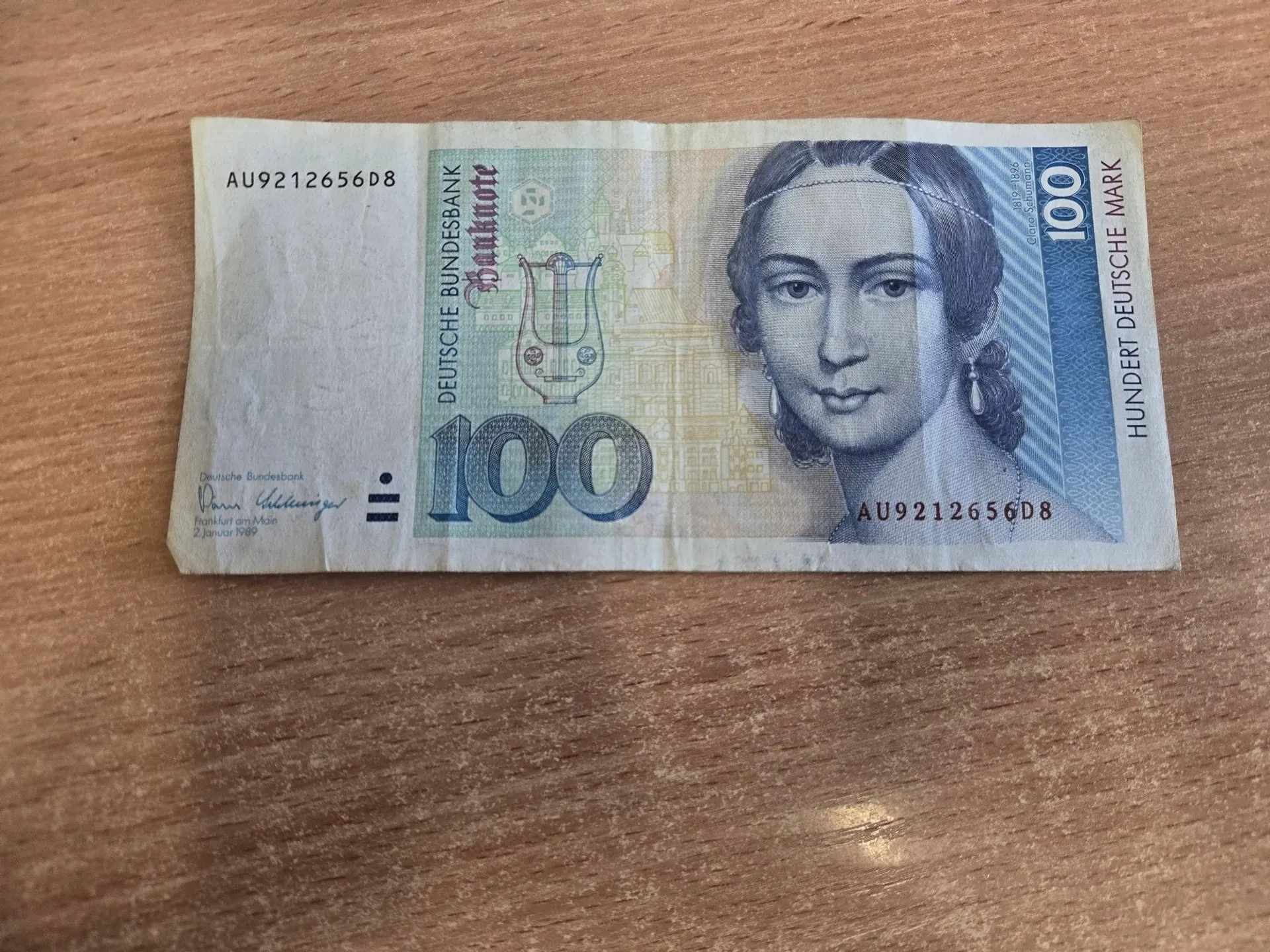 Niemiecki banknot o nominale 100 marek (Hundert Deutsche Mark). Na banknocie widnieje portret kobiety w ozdobnej opasce na głowie, liczba 100 oraz napisy: 'DEUTSCHE BUNDESBANK' i 'HUNDERT DEUTSCHE MARK'. Widoczny jest numer seryjny AU9212656D8. Banknot leży na drewnianej powierzchni.