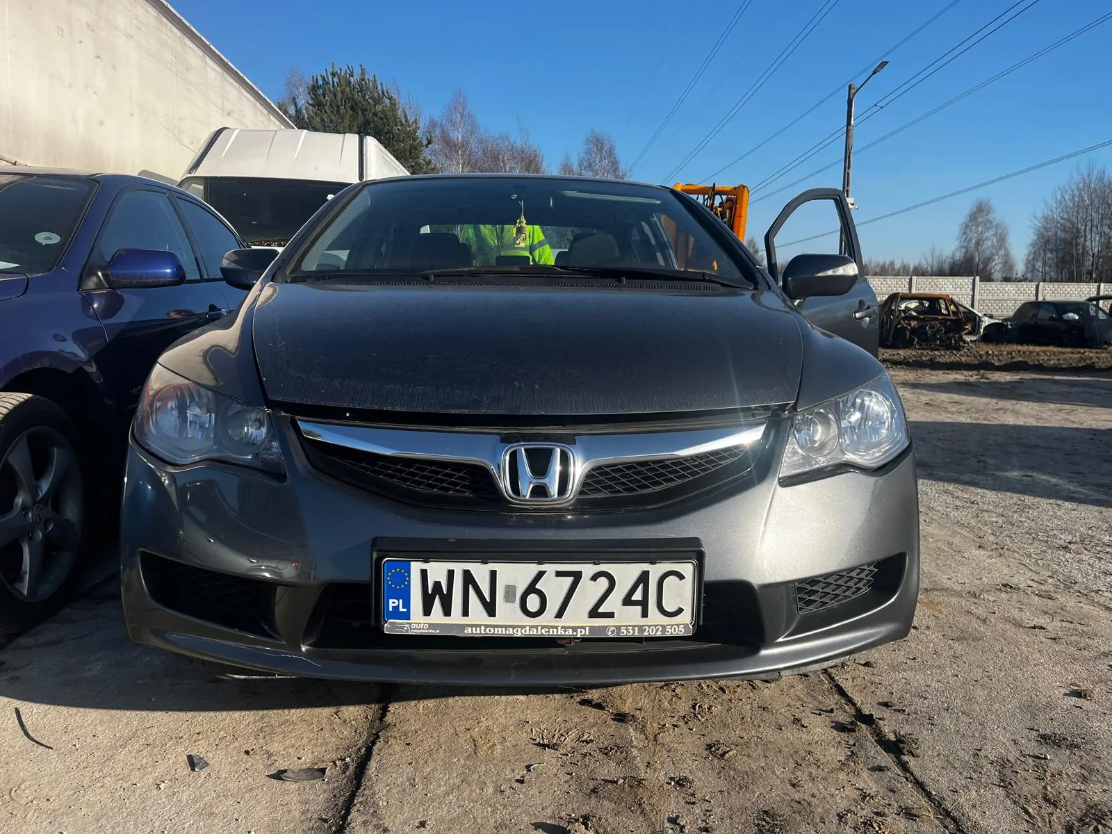 Przedni widok szarego samochodu marki Honda z tablicą rejestracyjną WN 6724C (PL, symbol Unii Europejskiej). Na tablicy widoczny napis 'automagdalenka.pl' oraz numer telefonu '531 202 505'. Samochód ma otwarte drzwi kierowcy i maskę. W tle: niebieski samochód po lewej, spalony pojazd, betonowa ściana, drzewa, słup z liniami energetycznymi oraz osoba w zielonej odzieży odblaskowej siedząca wewnątrz Hondy. Niebo jest bezchmurne, a podłoga asfaltowa z widocznymi pęknięciami.