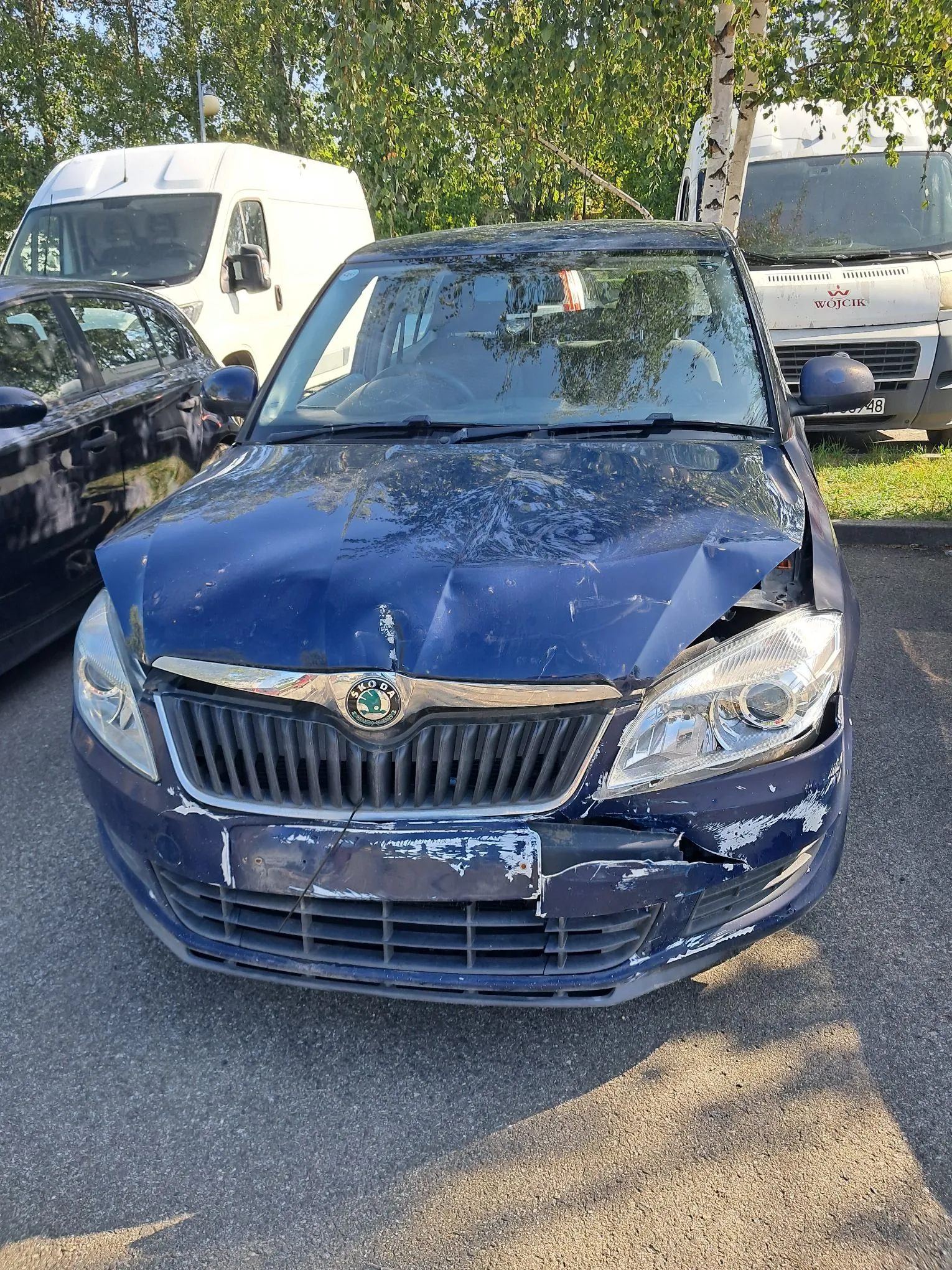 Niebieski samochód osobowy marki Skoda z poważnymi uszkodzeniami przodu. Maska jest mocno wgnieciona i pognieciona, a przedni zderzak jest pęknięty i częściowo oderwany. W tle widoczne są dwa białe samochody dostawcze oraz zielone drzewa.