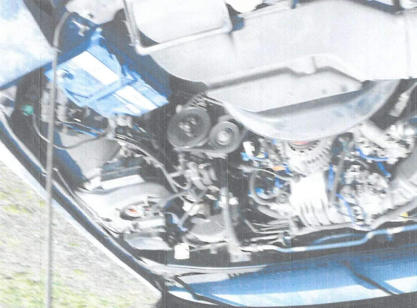 Zdjęcie przedstawiające komorę silnika samochodu z widocznymi elementami mechanicznymi, przewodami oraz niebieskim akumulatorem w górnej części kadru. Obraz jest wykonany pod kątem, co sprawia, że elementy silnika są ustawione pionowo zamiast poziomo.