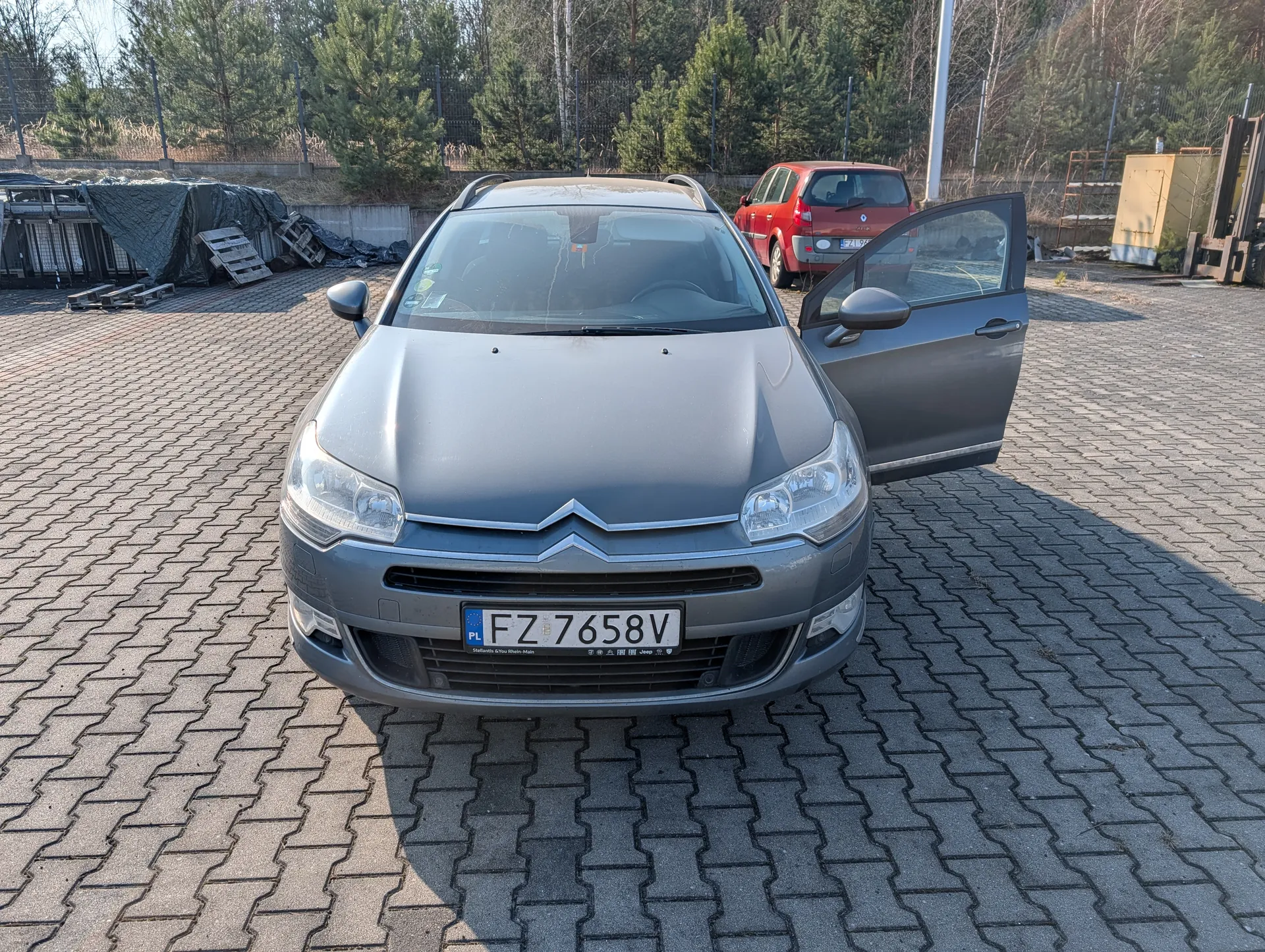 Samochód marki Citroën w kolorze szarym z otwartą drzwiami kierowcy, z tablicą rejestracyjną PL FZ 7658V. W tle czerwony samochód, drzewa, ogrodzenie oraz zakryte przedmioty na podwórku z bruku.