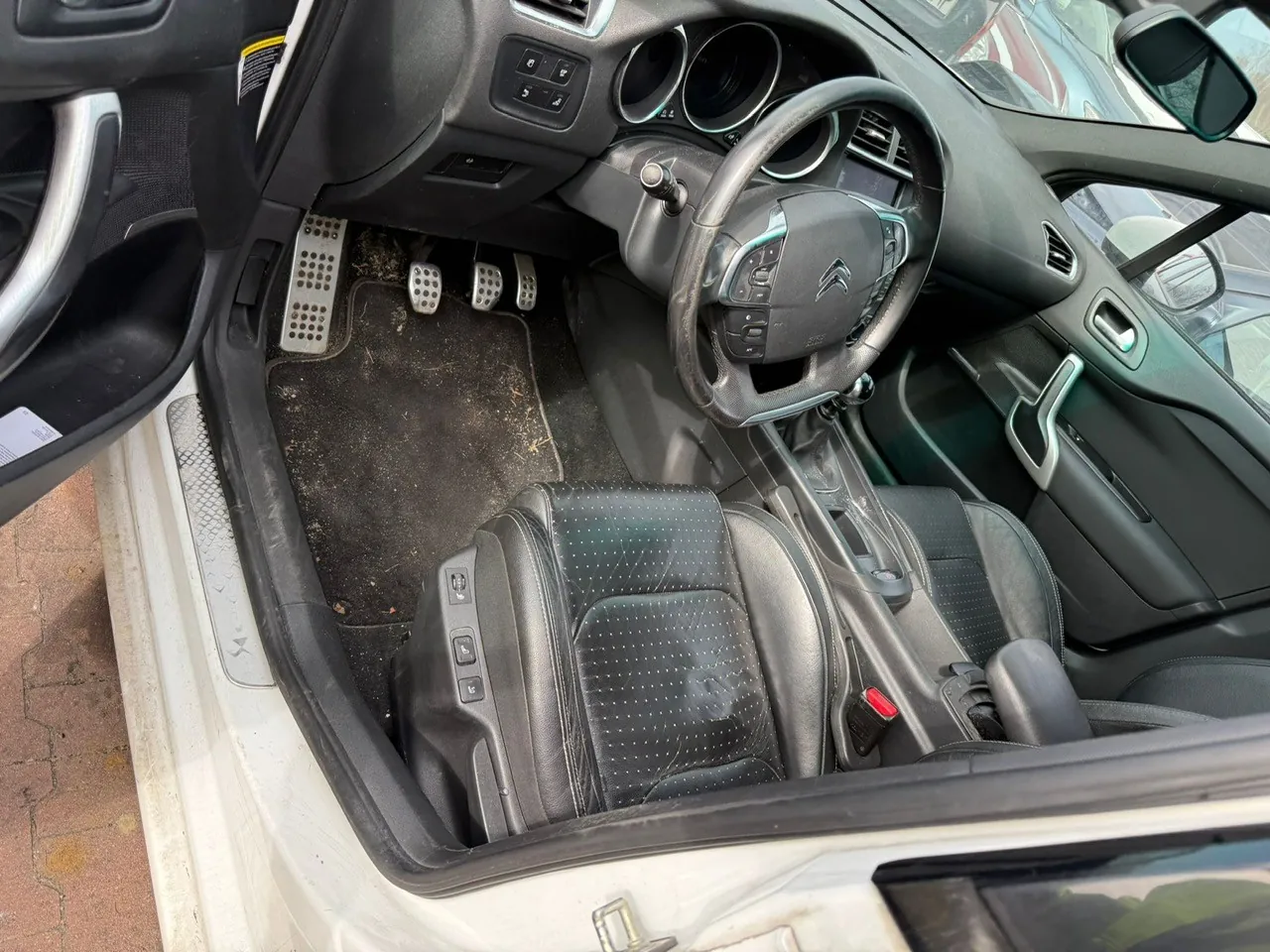 Wnętrze samochodu z otwartą lewą przodem drzwiami. Widoczna czarna kierownica z logo Citroën, czarne skórzane fotele z perforacją, trzy pedały (gaz, hamulec, sprzęgło), brudne dywany podłogowe, deska rozdzielcza z okrągłymi wydechami i przyciskami, dźwignia zmiany biegów oraz hamulec ręczny. Zewnętrzna część drzwi i nadwozie są białe. W tle fragment chodnika z czerwonych płytek.