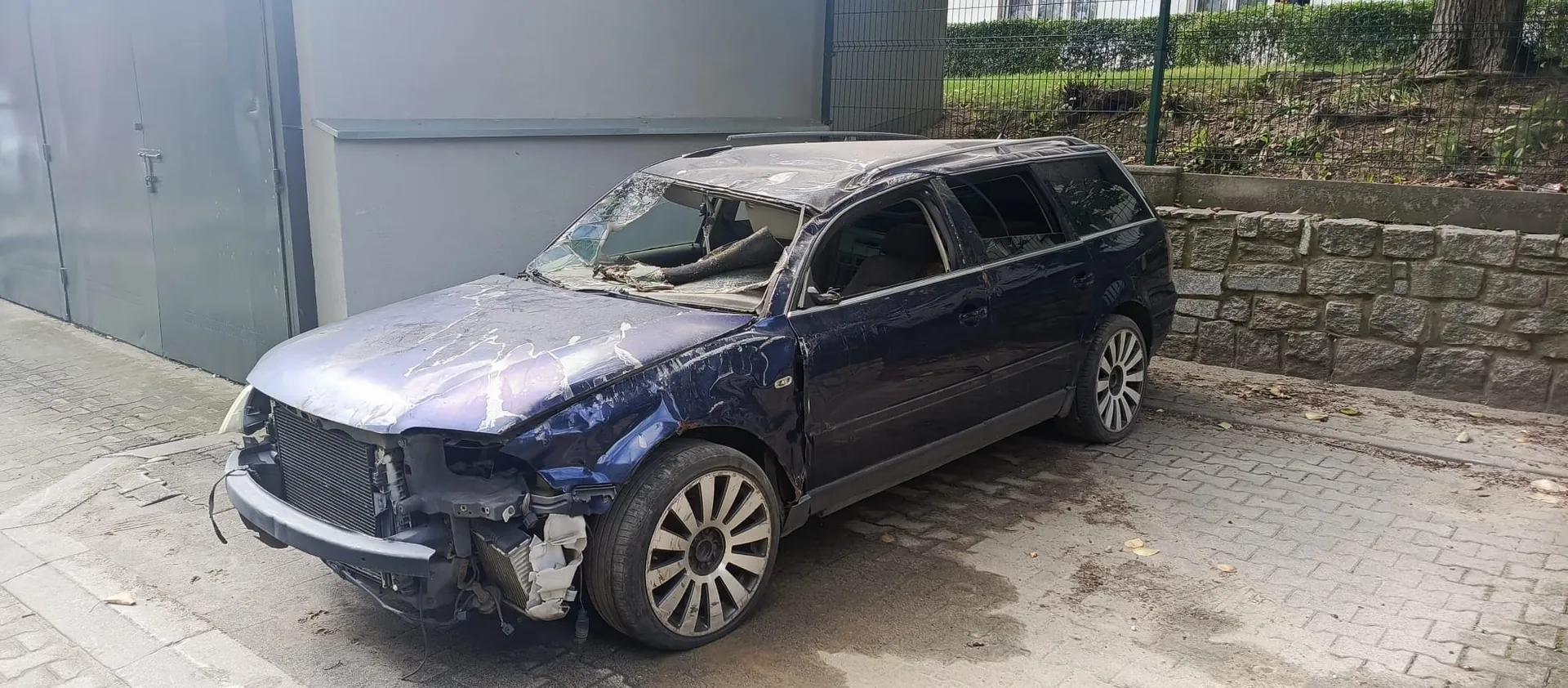 Ciemnoniebieski samochód typu kombi z poważnymi uszkodzeniami karoserii, zaparkowany na kostce brukowej obok szarej ściany i kamiennego murka. Pojazd ma zmiażdżony dach, brak przedniego zderzaka oraz uszkodzony przód i maskę. Szyby są wybite.