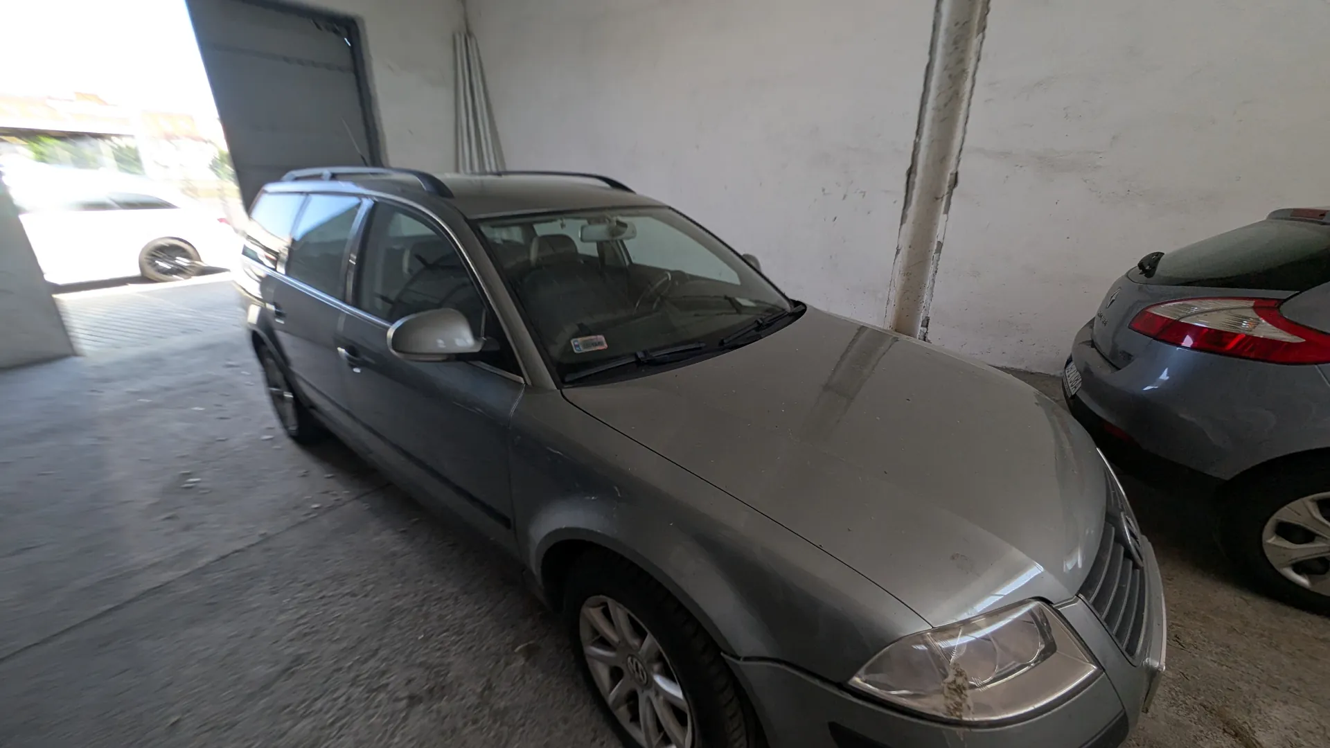 Srebrny samochód typu kombi marki Volkswagen zaparkowany w garażu. Obok niego stoi szary samochód osobowy. W tle otwarte drzwi garażu z widocznym białym samochodem na zewnątrz. Podłoga z betonu, białe ściany. Na przedniej szybie srebrnego samochodu widoczny mały naklejony znaczek.