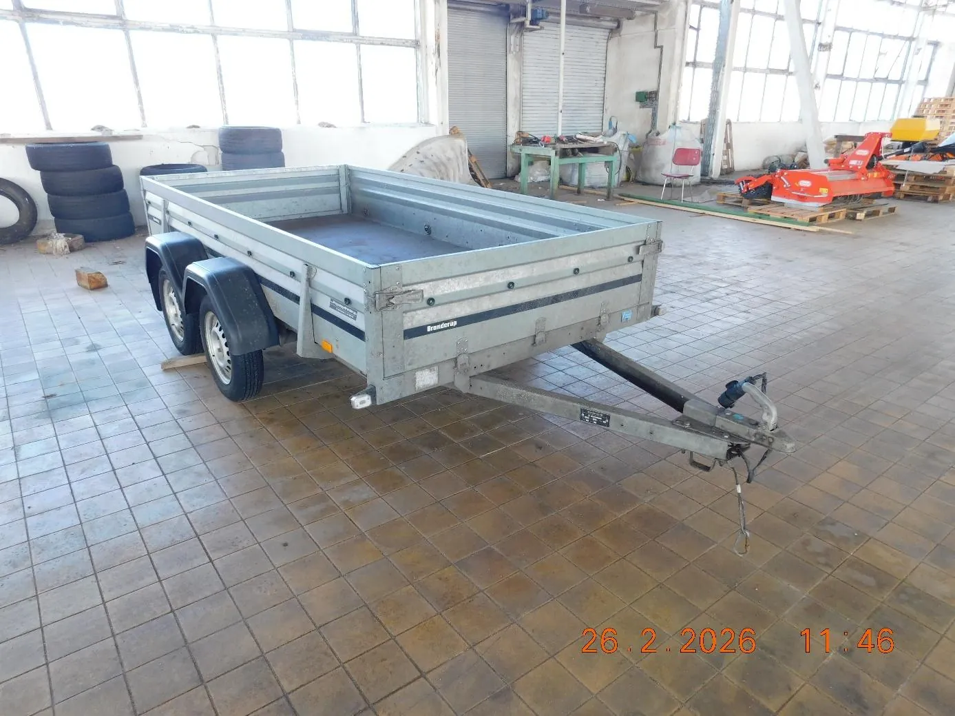 Srebrny przyczepa ciężarowa marki Brenderup z dwoma osiami i hakami do holowania, umieszczona w przestronnym warsztacie z podłogą z brązowych płytek. W tle widoczne są stosy opon, zielony stół z narzędziami, czerwone krzesła, pomarańczowe maszyny rolnicze na paletach oraz duże okna. W prawym dolnym rogu obrazu znajduje się pomarańczowy znaczek z datą 26.2.2026 i godziną 11:46.