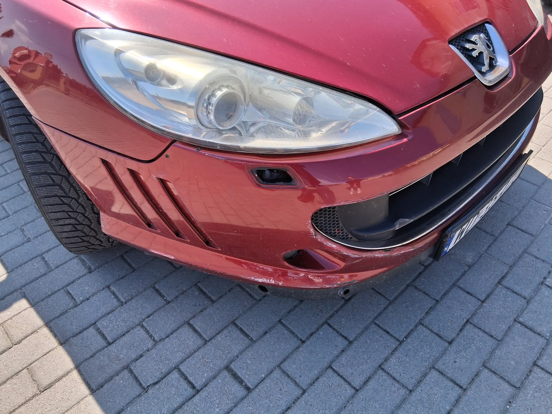 Przednia część czerwonego samochodu marki Peugeot z widocznym reflektorem, emblematem lwa na masce, czarnym zderzakiem z otworami wentylacyjnymi oraz częścią opony. Samochód stoi na brukowanej powierzchni z szarych płytek. Widoczna jest również fragmentaryczna tablica rejestracyjna po prawej stronie.