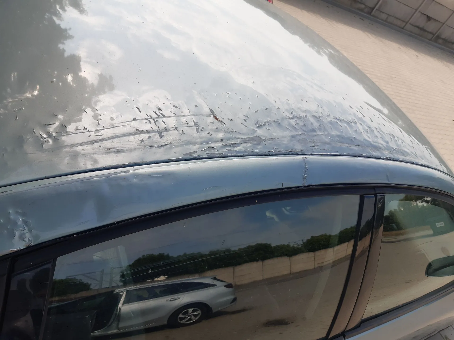 Zbliżenie na dach i górną krawędź bocznej szyby srebrno-niebieskiego samochodu. Lakier na dachu jest mocno uszkodzony, widoczne jest łuszczenie się i odwarstwianie warstwy lakieru. W szybie samochodu odbija się inne, srebrne auto typu kombi oraz otoczenie z zielenią i jasnym murem.