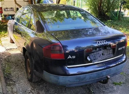 Tylny widok ciemnoniebieskiego samochodu marki Audi A6. Na klapie bagażnika widoczne jest logo Audi, oznaczenie modelu 'A6' oraz oznaczenie silnika '2.5 TDI'. Samochód nie posiada tablicy rejestracyjnej, a w miejscu jej montażu widoczna jest metalowa ramka. Pojazd posiada hak holowniczy. Auto zaparkowane jest na żwirowym podłożu obok zieleni.