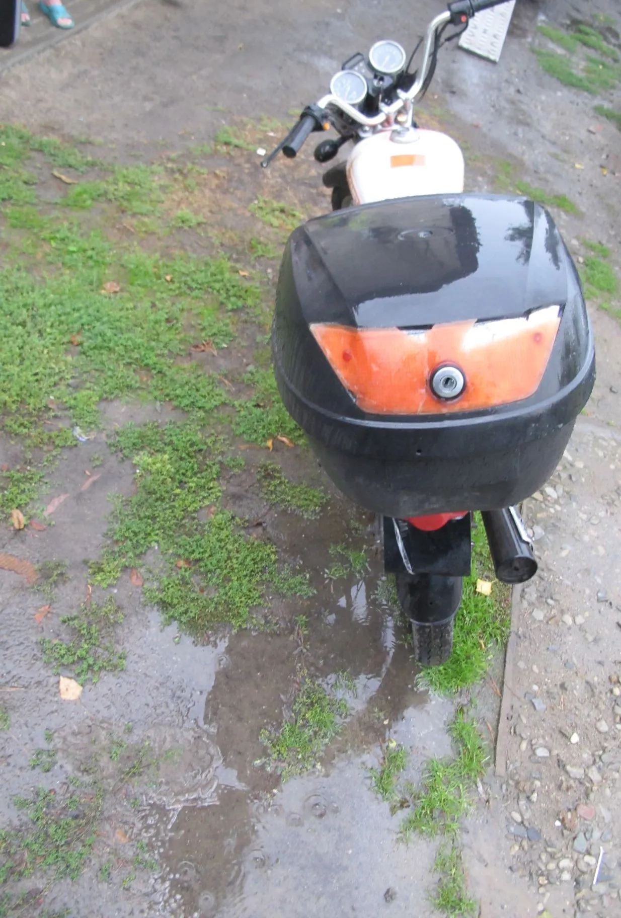 Czarny motocykl z pomarańczowym elementem na przedniej obudowie, zaparkowany na błotnistym podłożu z kępami zielonej trawy.