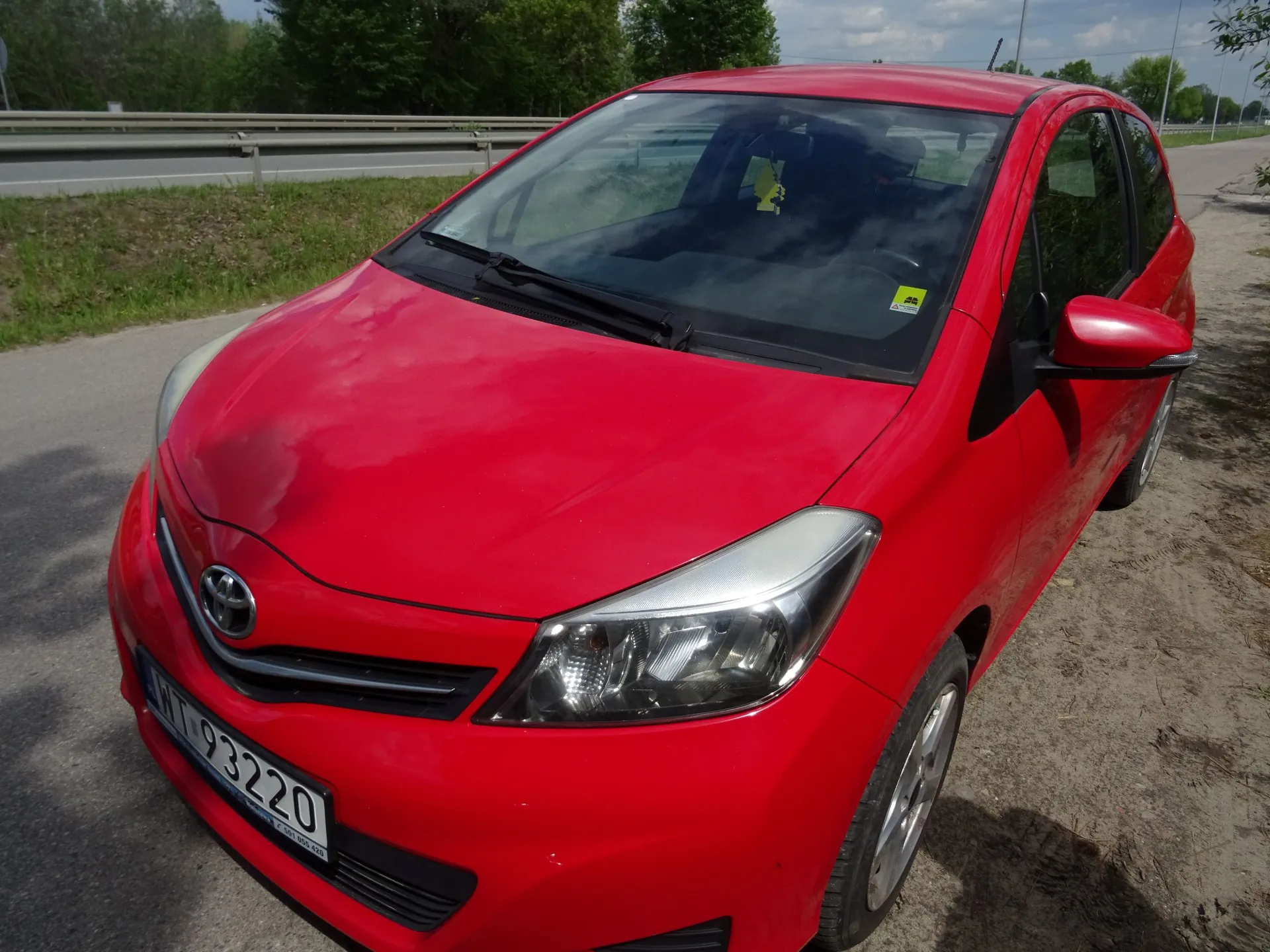 Czerwony samochód marki Toyota zaparkowany na poboczu drogi. Widoczny jest przód pojazdu z tablicą rejestracyjną o numerze WT 93220. W tle znajduje się barierka energochłonna, trawa oraz zielone drzewa pod jasnym niebem.