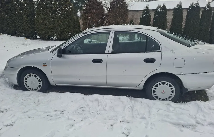 Srebrny samochód osobowy typu sedan zaparkowany na śniegu. Pojazd ma cztery drzwi, na felgach widoczny jest logo Mitsubishi. Na dachu i masku samochodu leży niewielka warstwa śniegu. W tle rosną zasłonięte śniegiem krzewy i drzewa iglaste, a za nimi budynek z dachem pokrytym śniegiem. Śnieg pokrywa również całą powierzchnię ziemi wokół pojazdu, z widocznymi odciskami stóp.