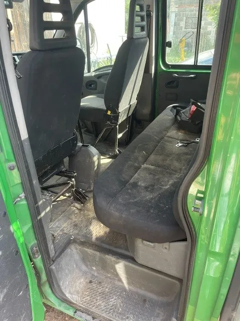 Wnętrze kabiny zielonego samochodu dostawczego. Widoczne są dwa przednie fotele z czarnej tkaniny oraz tylna kanapa w kolorze czarnym, która jest zakurzona. Na kanapie leży czarna torba. Podłoga jest wykonana z szarego tworzywa sztucznego i jest zabrudzona pyłem. Widoczny jest fragment zielonego nadwozia oraz otwarta boczna drzwi.