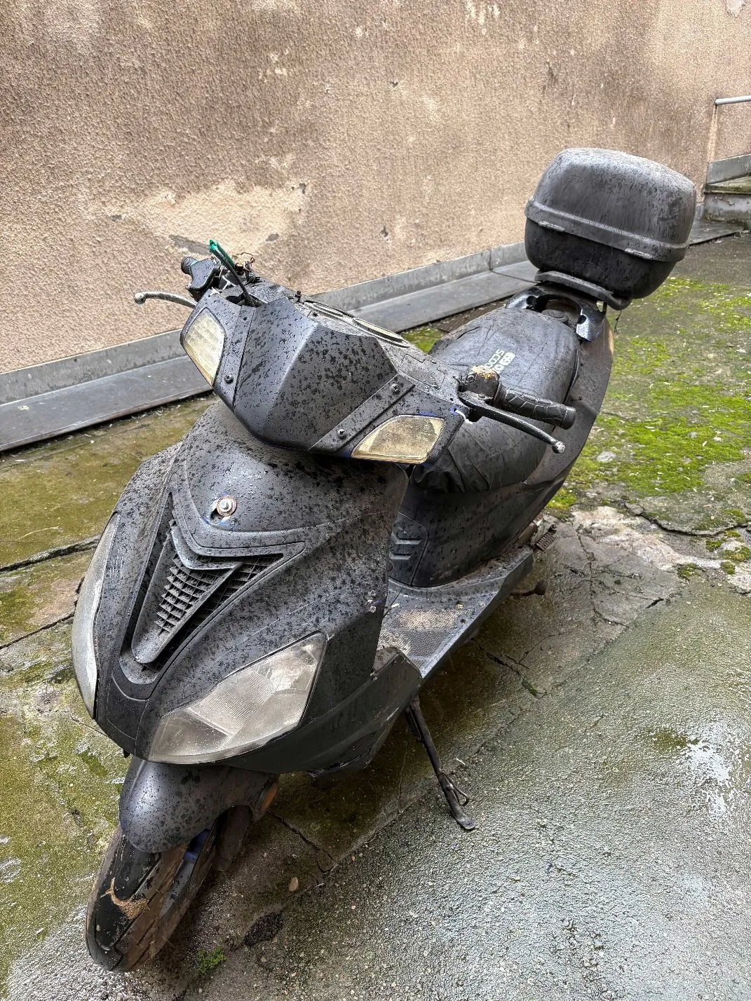 Brudny, czarny skuter zaparkowany na betonowym podłożu obok ściany budynku. Pojazd jest pokryty warstwą kurzu i błota, posiada zamontowany czarny kufer z tyłu. Skuter stoi na bocznej stopce.