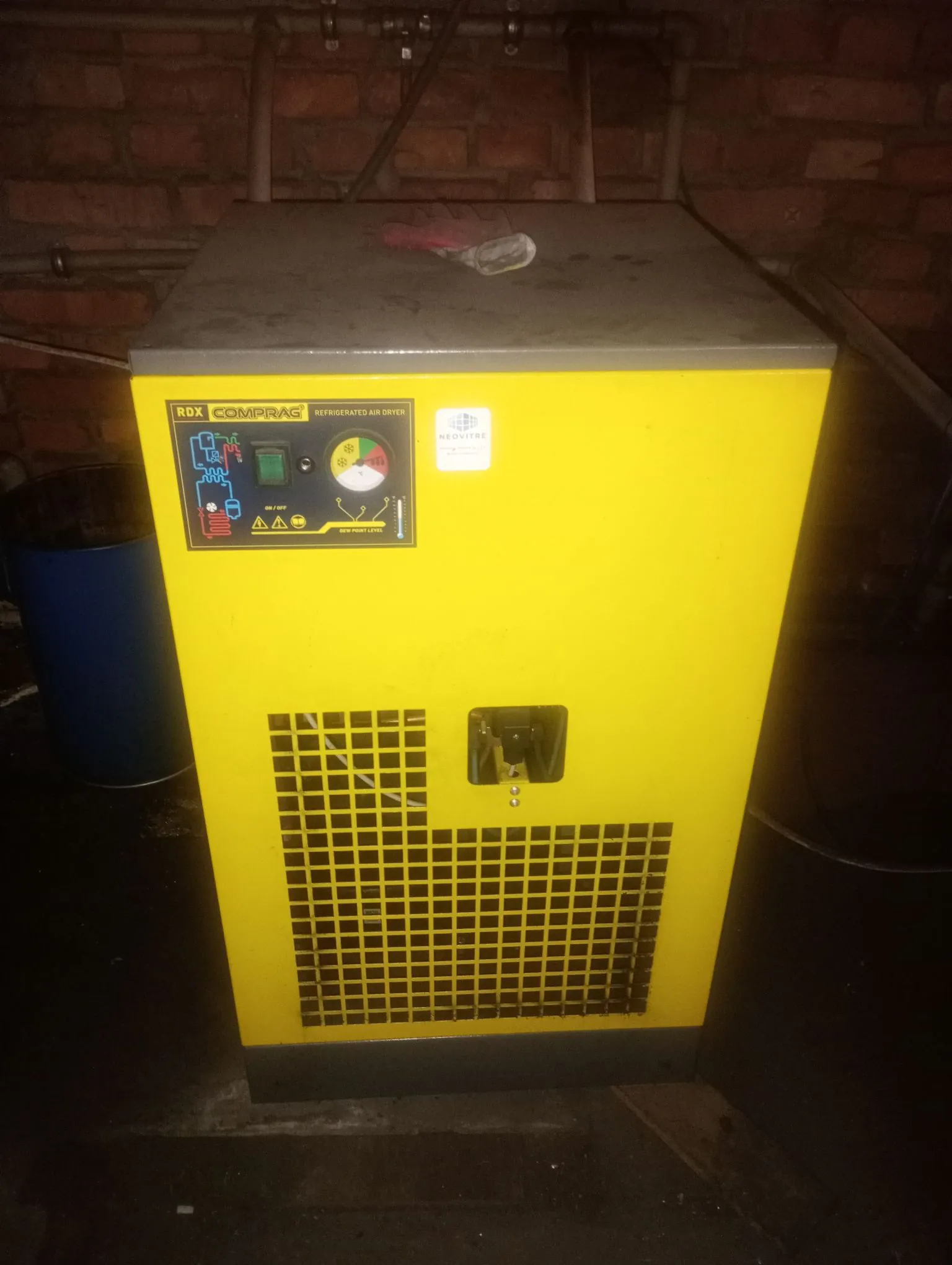 Żółta, metalowa maszyna przemysłowa, osuszacz powietrza marki COMPRAG model RDX Refrigerated Air Dryer. Urządzenie posiada panel sterowania z przyciskami, wskaźnikami i tekstem, a w dolnej części znajduje się czarna kratka wentylacyjna. Maszyna stoi na ciemnym podłożu na tle czerwonej ceglanej ściany. Po lewej stronie widoczny jest fragment niebieskiego pojemnika.